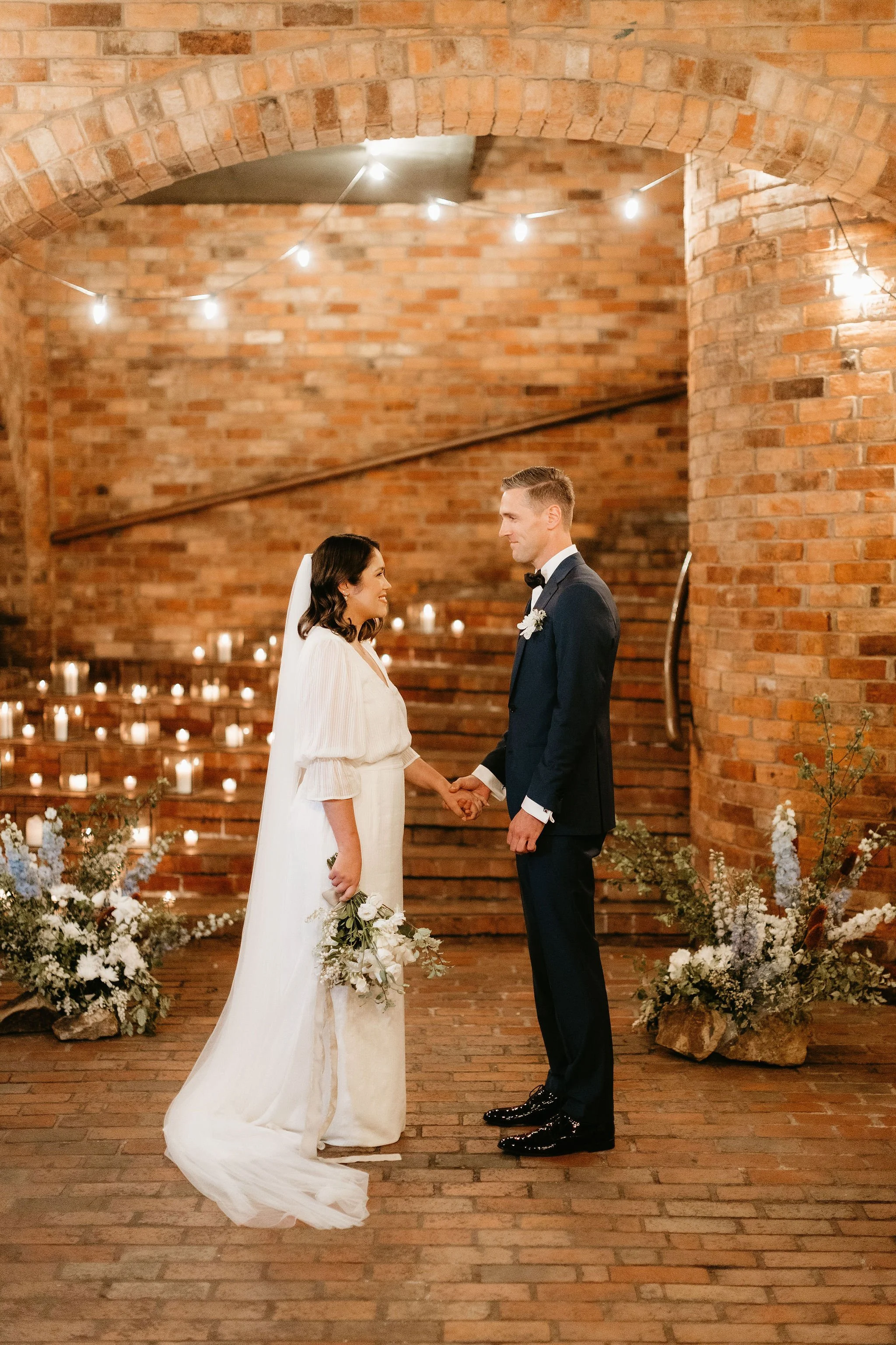 A bride and groom stand hand in hand facing each other during their wedding ceremony in a warmly lit, rustic brick venue. The bride wears a flowing white gown and holds a bouquet, while the groom wears a dark suit with a bow tie and boutonnière. The background features candles and floral arrangements.