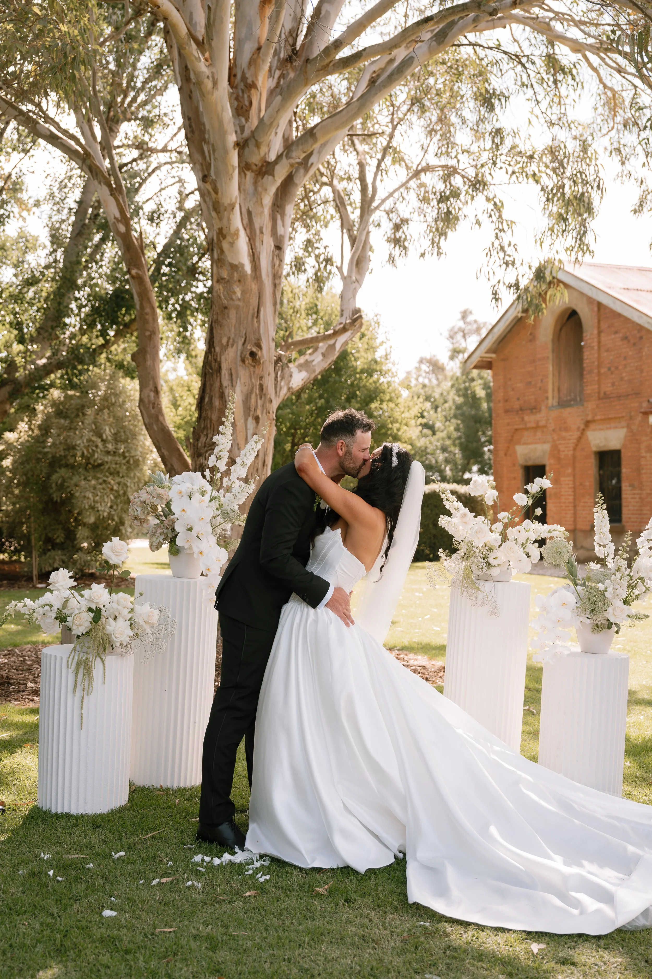 A newlywed couple shares a kiss during an outdoor wedding ceremony with floral arrangements and a large tree in the background.