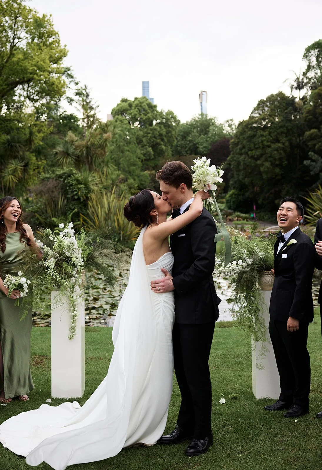 A wedding ceremony outdoors with a bride and groom kissing, surrounded by bridesmaids and groomsmen, greenery, and a pond in the background.