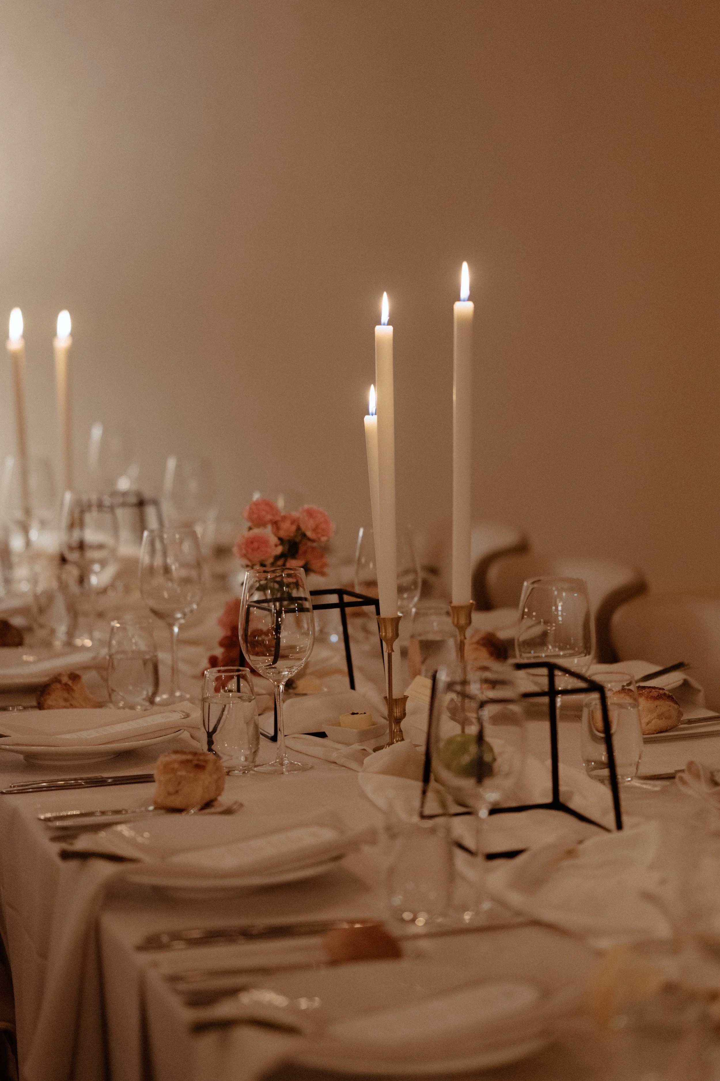 A formal dining table set with wine glasses, plates, silverware, and napkins, decorated with pink flowers and tall white candles with flames burning.