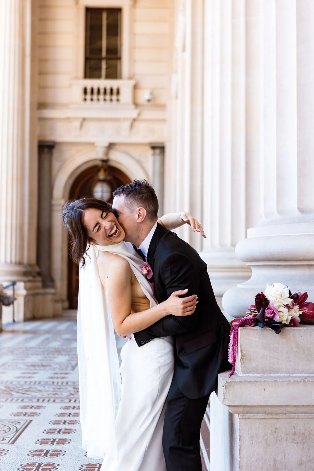 Two people, a man and a woman, sharing a joyful kiss during a wedding. The woman wears a white wedding dress, and the man wears a black tuxedo. They are standing near large white columns inside a grand building with ornate architectural details and a floral arrangement on a stone pedestal.