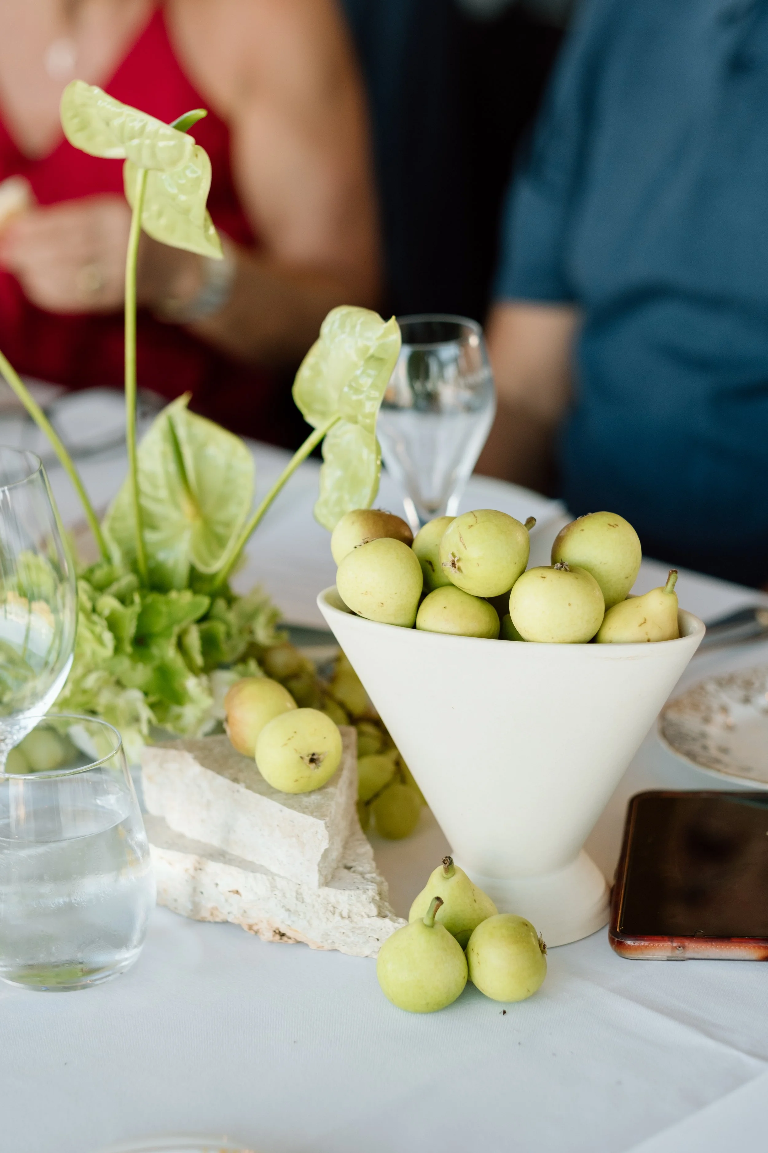 A white ceramic bowl filled with small, yellow-green apples on a table. There are also some apples scattered on the table. A glass of water, a smartphone, and some glassware are visible nearby. In the background, people are visible, one wearing a red