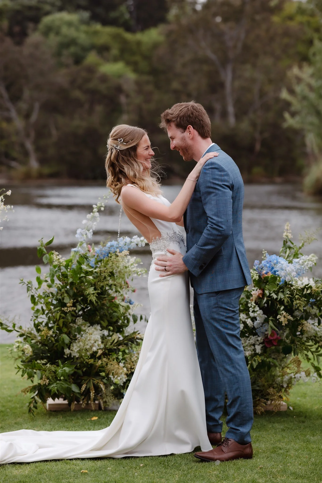 A bride and groom standing closely together outdoors by a river, looking at each other smiling. The bride is in a white wedding dress with a floral accent in her hair, and the groom is in a blue suit. The scene is decorated with flowers and greenery, with trees and a river in the background.