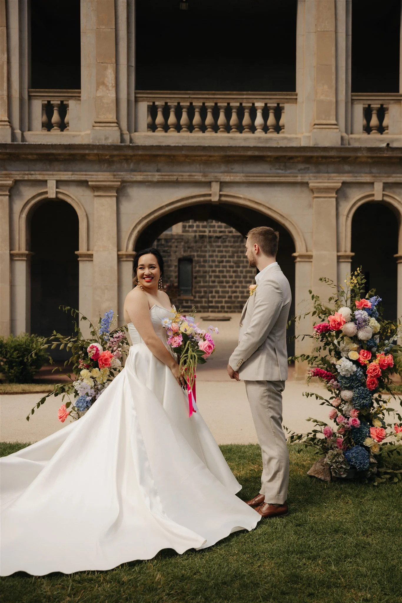 A bride and groom standing outdoors in front of a historical building with arches and columns. The bride is holding a bouquet of colorful flowers, smiling, and wearing a white wedding gown. The groom is looking at her, dressed in a light-colored suit. There are large floral arrangements on either side of the couple.