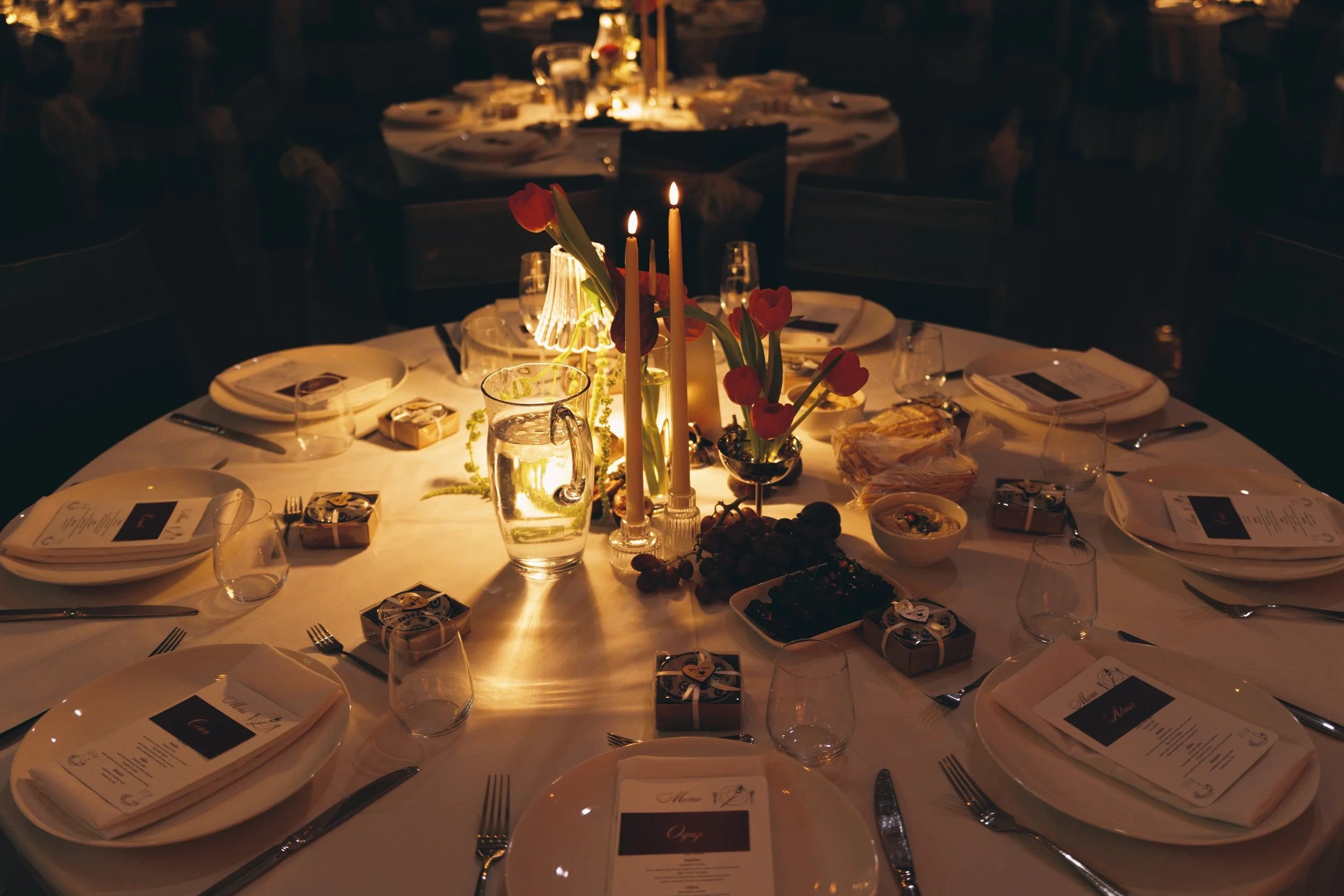 A round table set for a formal dinner with white tablecloth, plates, silverware, wine glasses, and printed menus. The centerpiece has red tulips in a glass vase illuminated by candlelight, creating a warm glow in a dimly lit room.