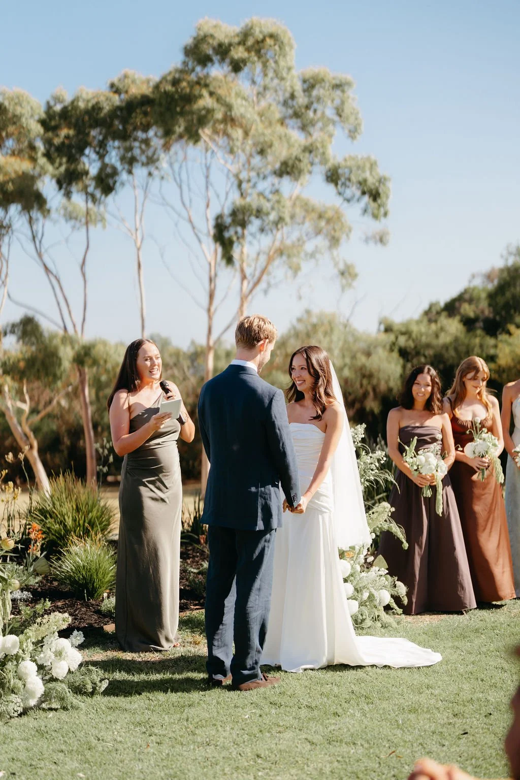 A couple getting married outdoors with an officiant and bridesmaids present, during a sunny day.