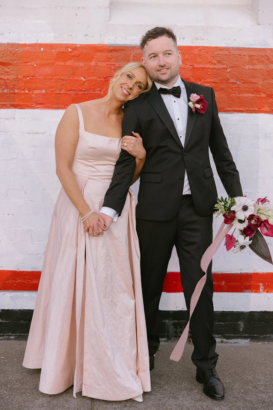 A bride and groom holding hands and smiling for a photo in front of a white brick wall with a red stripe. The bride wears a light pink strapless gown and the groom is in a black tuxedo with a bow tie and a red flower boutonniere. The bride is leaning her head on the groom’s shoulder and has a bouquet of flowers.