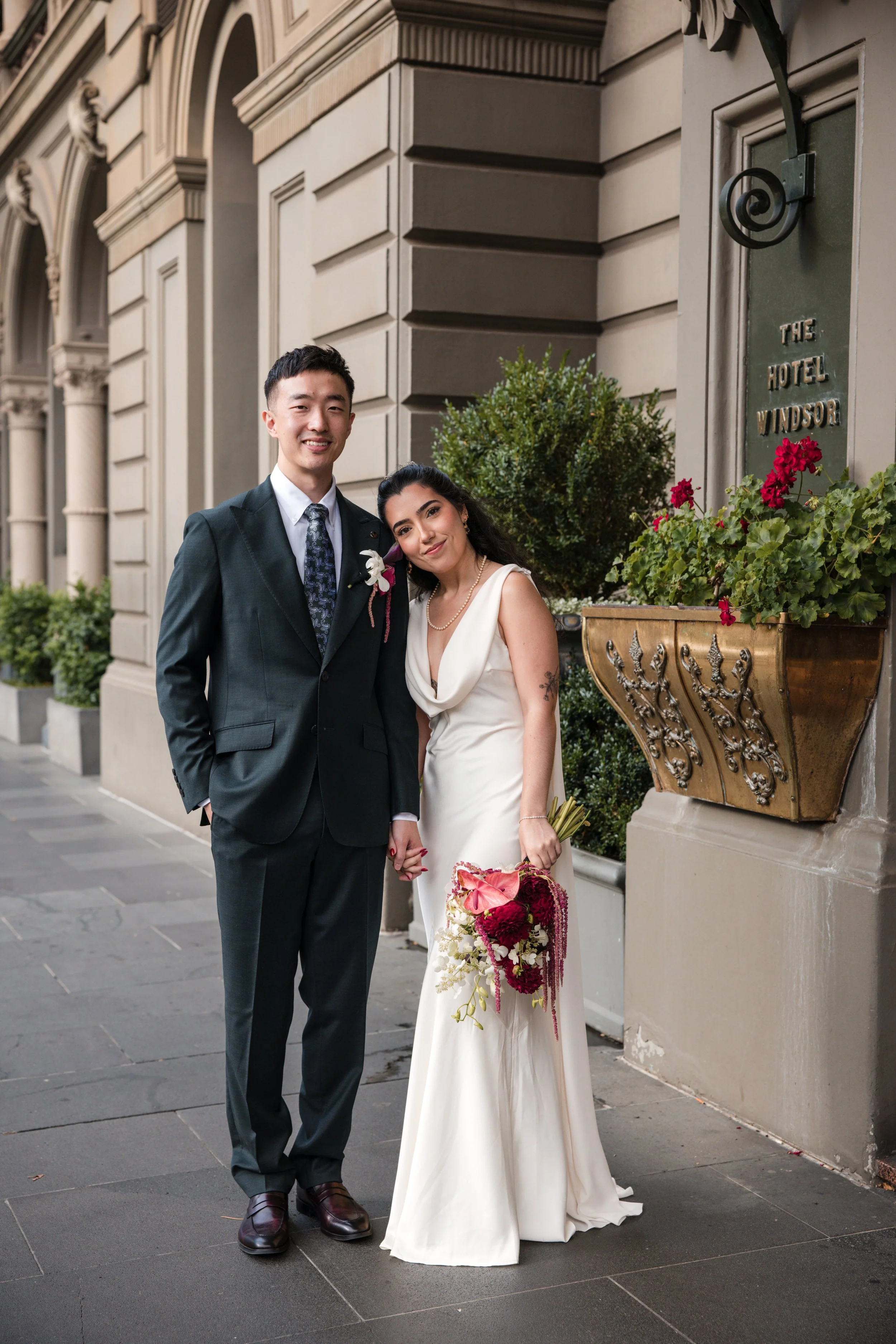 A happy couple in wedding attire standing outside a hotel, holding hands, with the man in a dark suit and the woman in a white gown holding a bouquet of flowers.