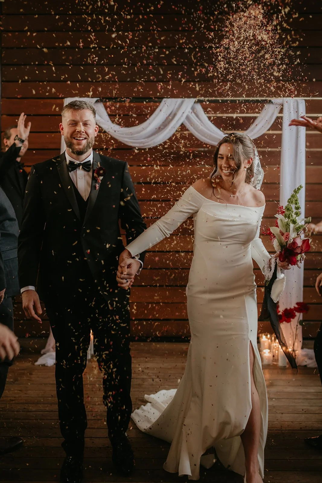 A bride and groom holding hands and smiling, surrounded by confetti during wedding celebration. The groom is dressed in a black tuxedo with a bow tie, and the bride is wearing an off-the-shoulder wedding dress and holding a bouquet of flowers. Wooden panel wall with white drapery in the background.