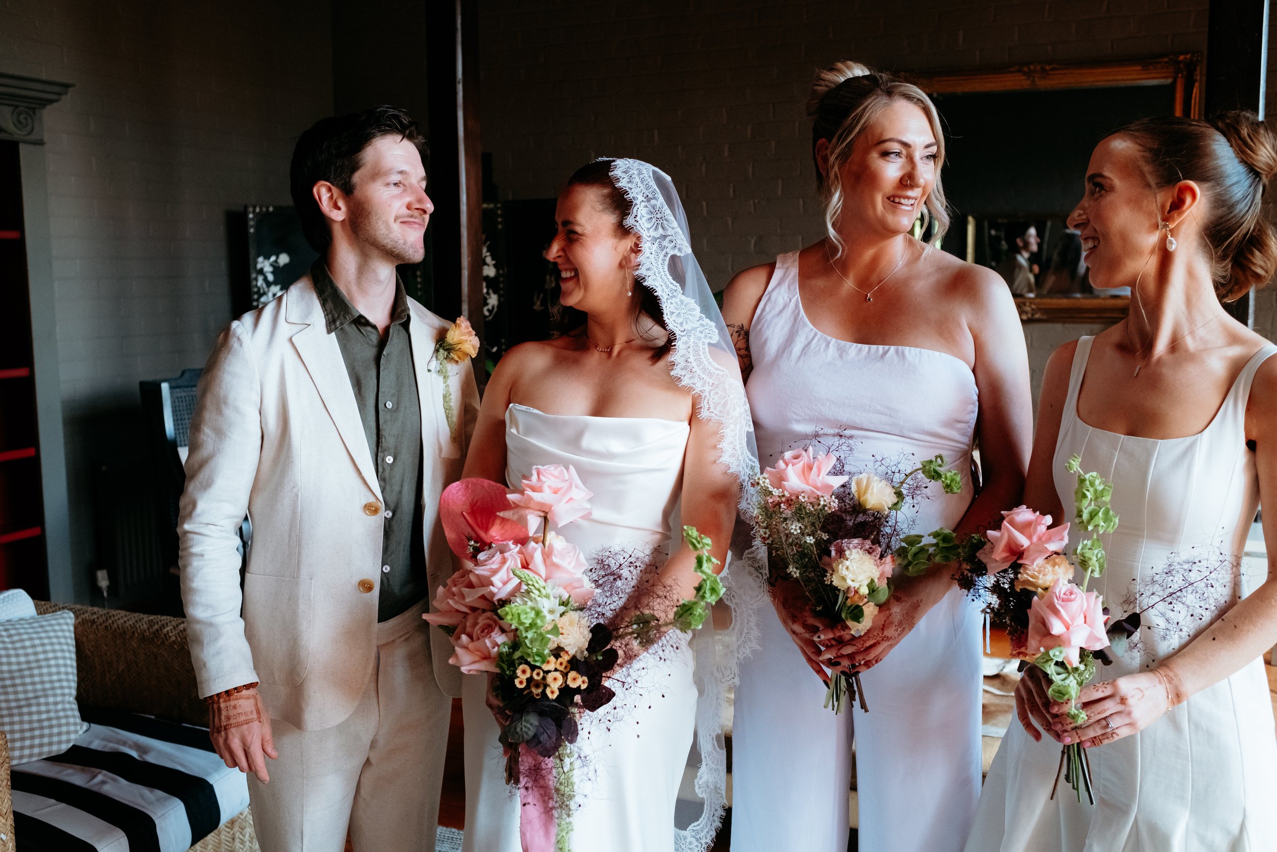 Group of people celebrating a wedding, with the bride and groom standing in the center, holding colorful bouquets, and smiling at each other, surrounded by bridesmaids and guests.