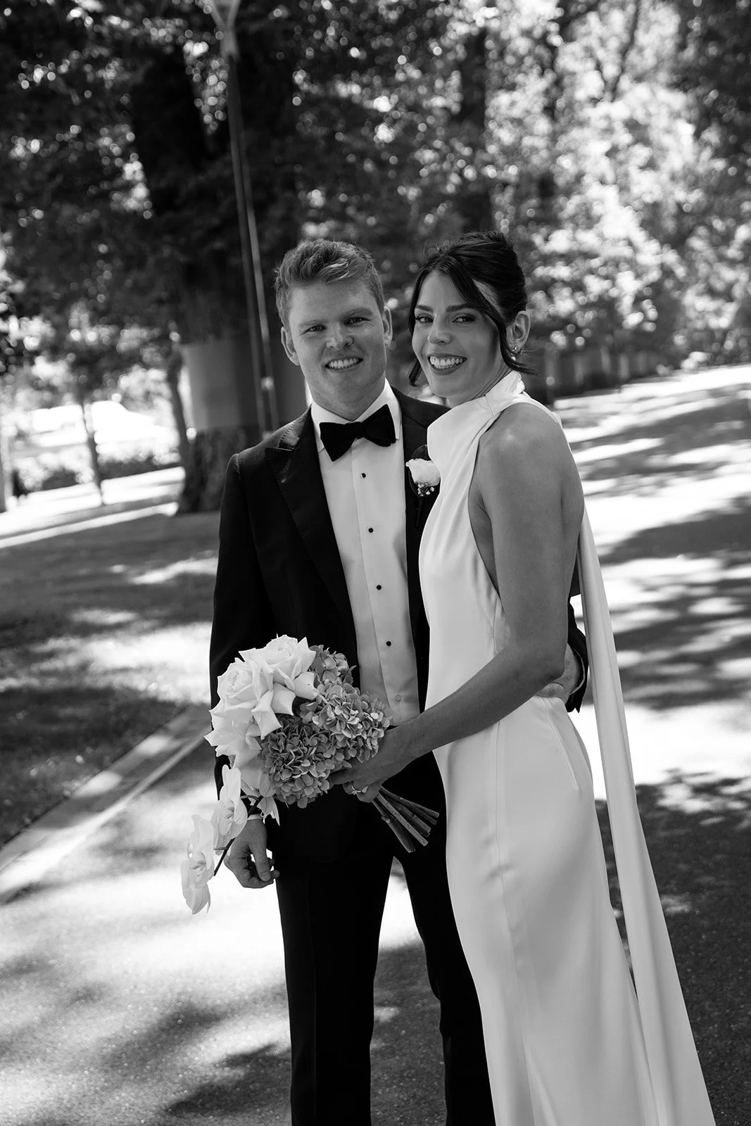 Black and white photo of a smiling bride and groom standing outdoors, holding a bouquet of flowers, surrounded by trees.