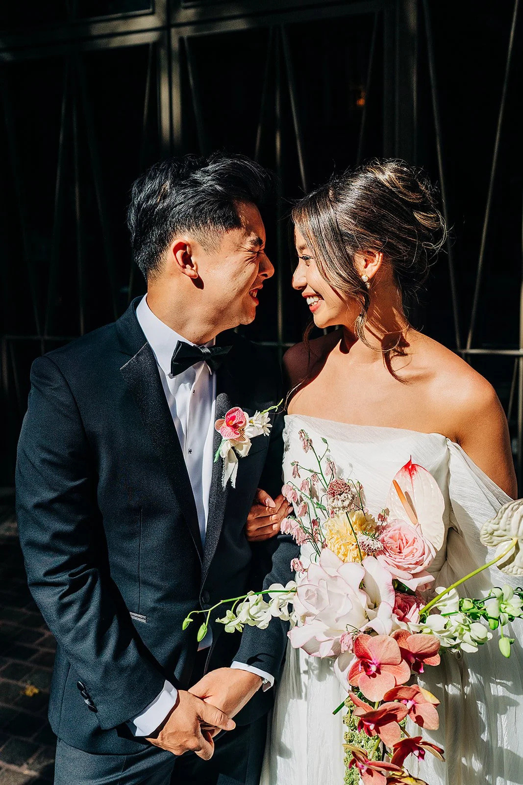 A couple in wedding attire standing close together, smiling and touching foreheads, with a floral bouquet in front of them.