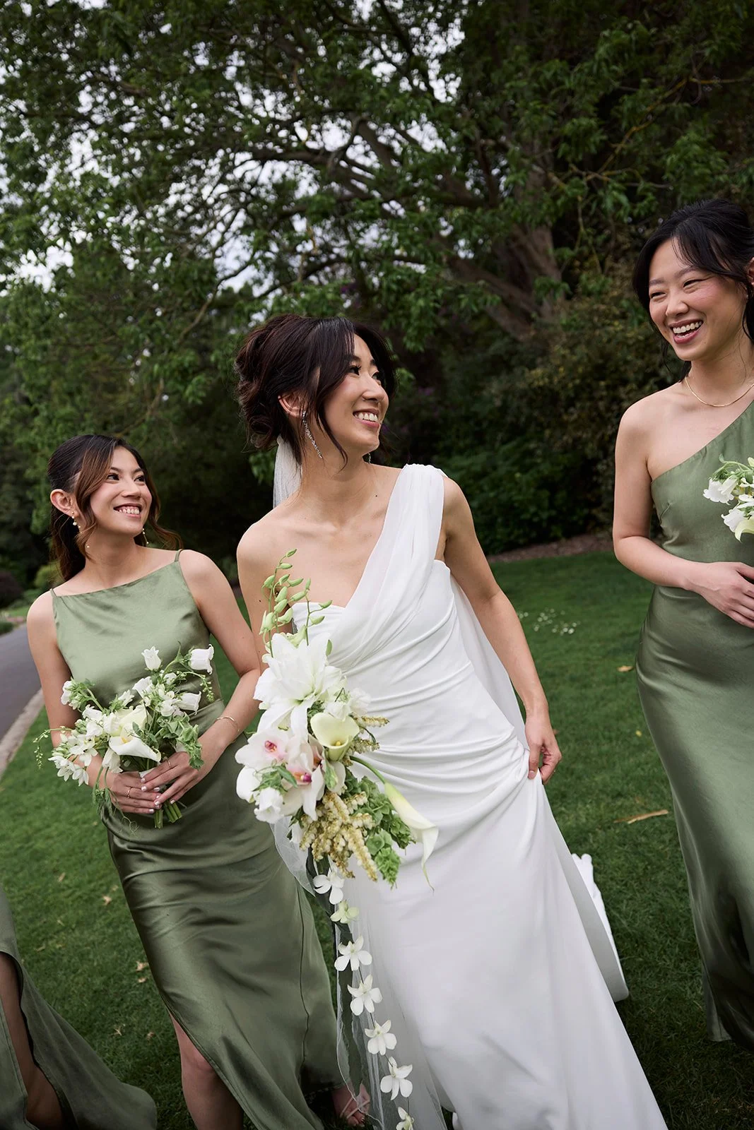 Three women, including a bride in a white wedding dress and two bridesmaids in green dresses, walking together outdoors with trees in the background, holding bouquets, smiling and enjoying the moment.