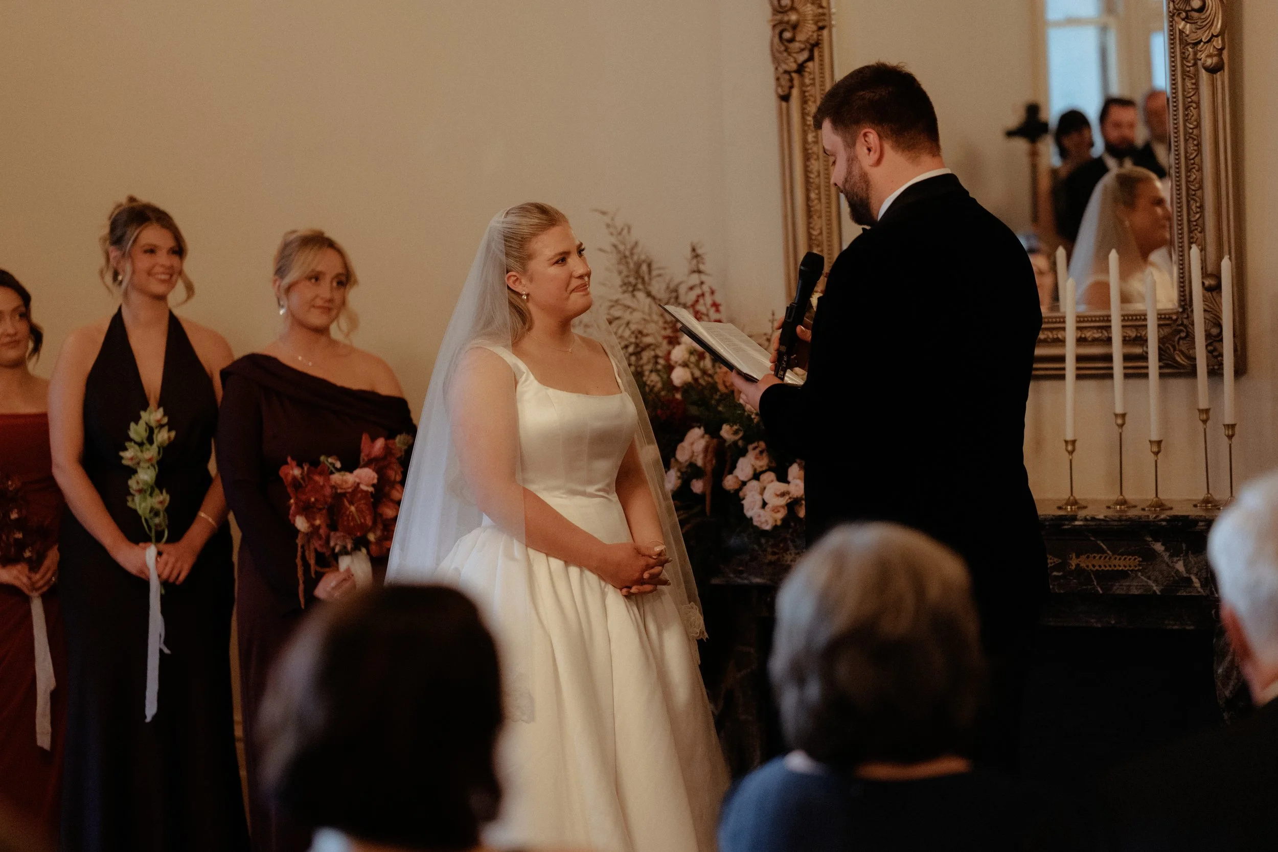 A bride and groom exchange vows during a wedding ceremony in a decorated indoor setting, with bridesmaids standing behind the bride and guests observing.