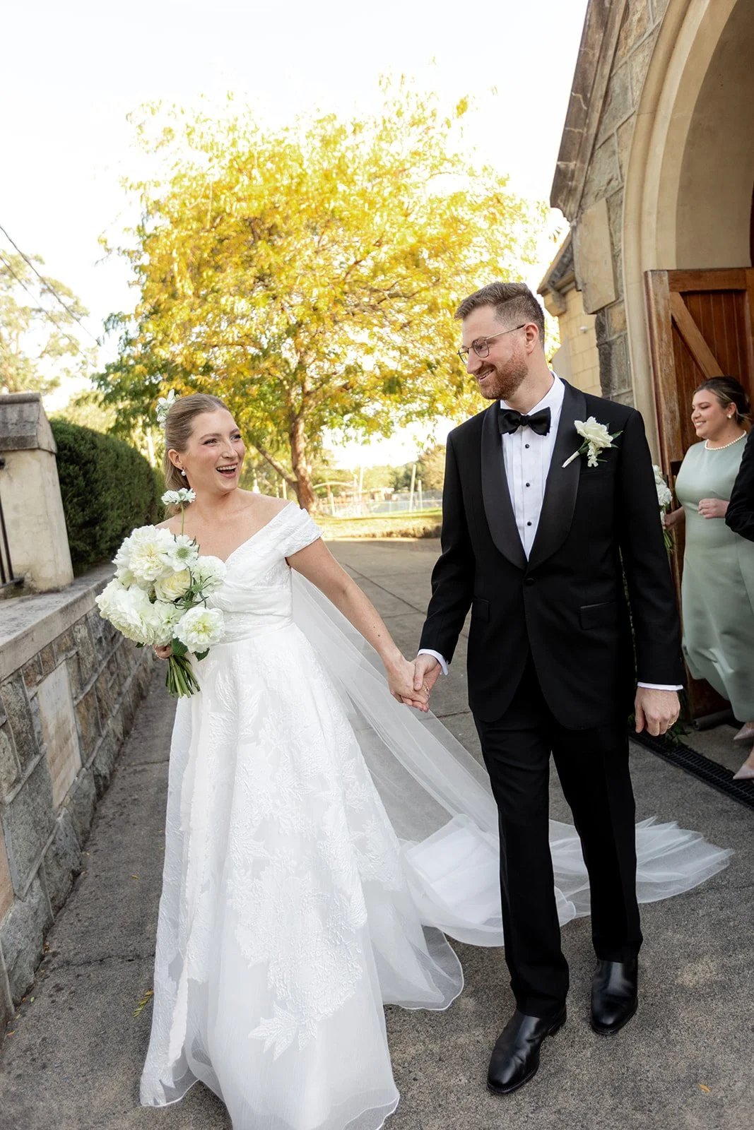 A bride and groom hold hands and smile at each other outside a church, with a bridesmaid in the background. The bride wears a white wedding dress and holds a bouquet of white flowers, while the groom wears a black tuxedo with a bow tie and a white boutonniere. The scene is set outdoors with trees and a stone wall, and the weather appears clear and sunny.