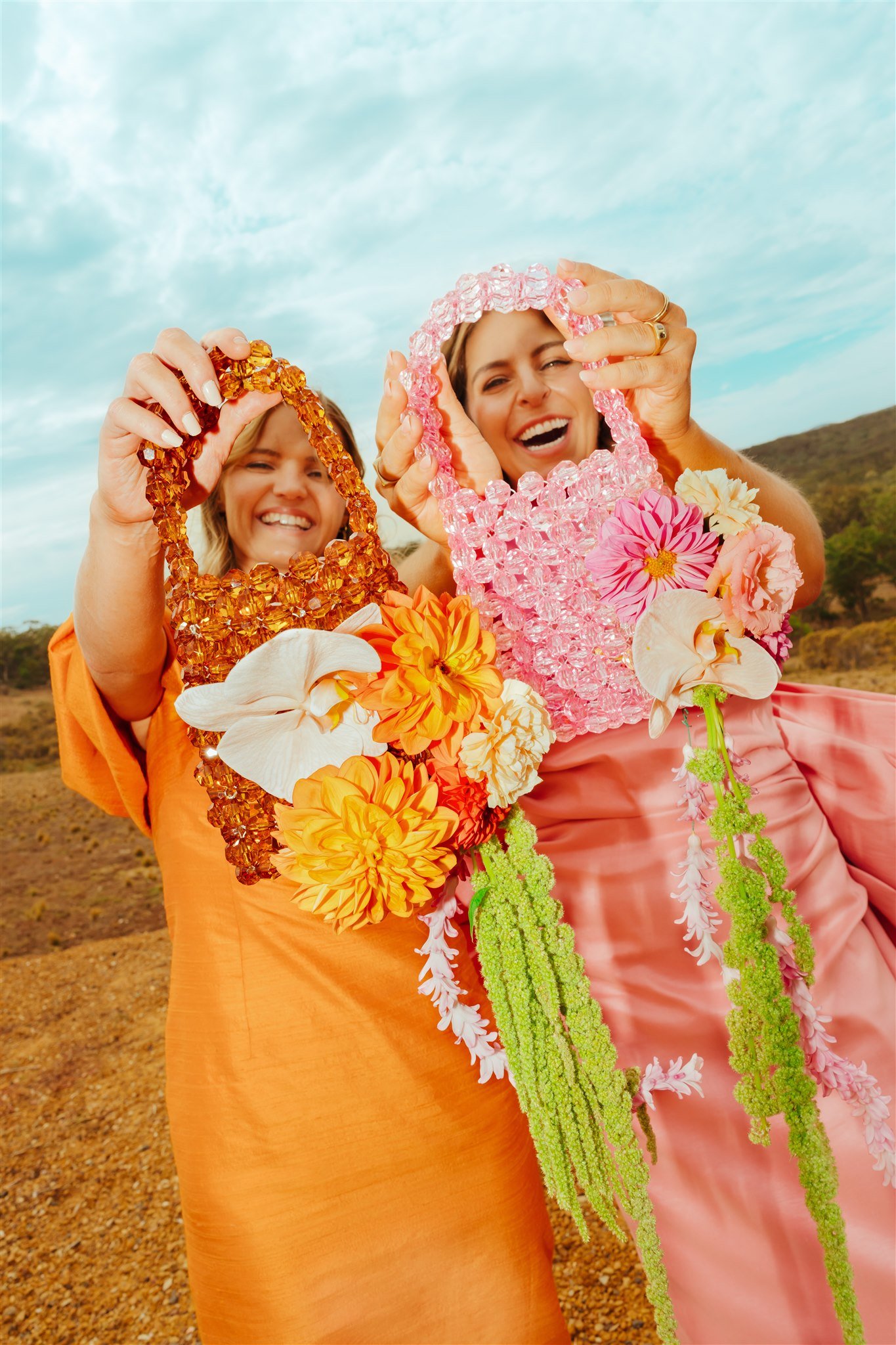 Two smiling women holding floral and beaded necklaces outdoors with a landscape background.