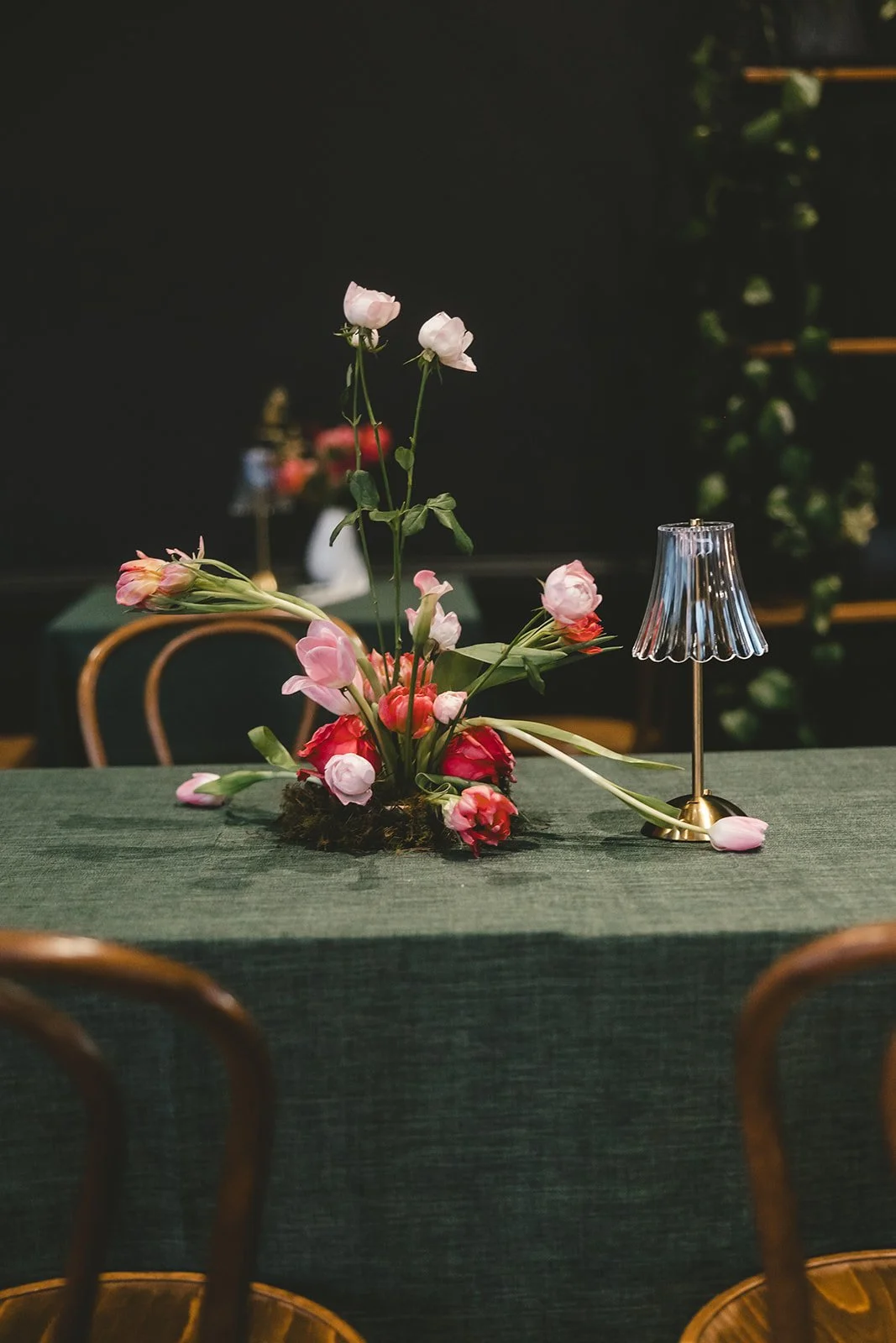 A floral centerpiece of pink and red flowers on a green tablecloth, accompanied by a small gold lamp with a glass shade in a dark room.