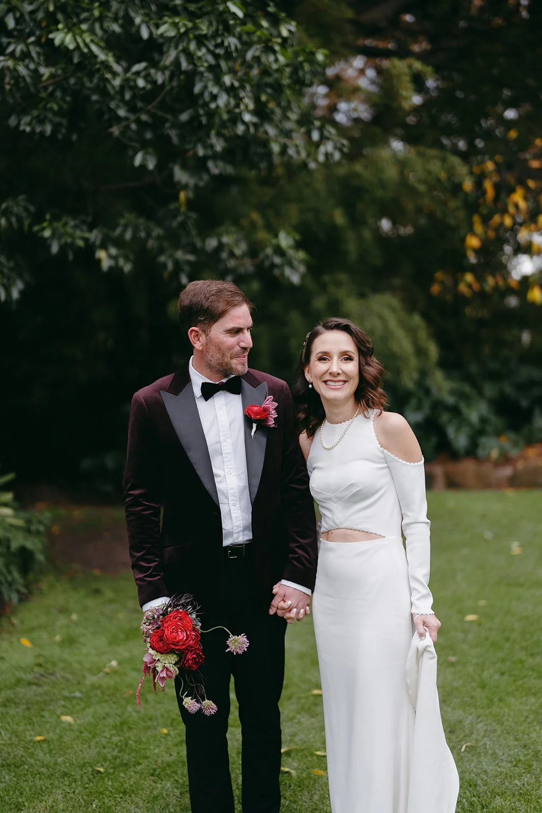 A newlywed couple holding hands on a grassy garden with trees in the background. The groom wearing a black tuxedo and bow tie, holding a bouquet of red flowers. The bride in a white dress with cutouts, smiling. Both appear happy.