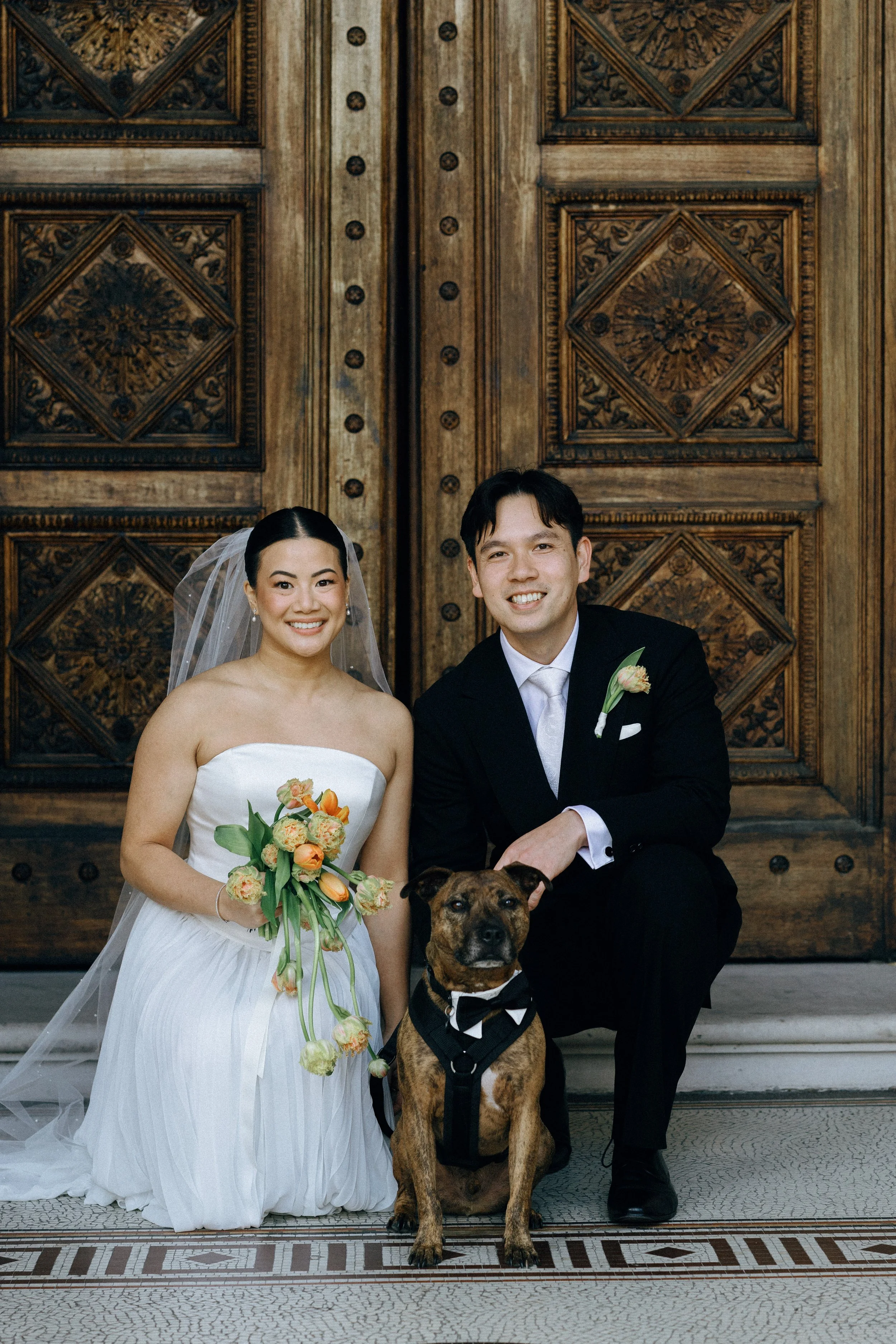 A bride and groom posing with a brown dog dressed in a tuxedo, standing in front of a wooden door with elaborate carvings, during a wedding celebration.