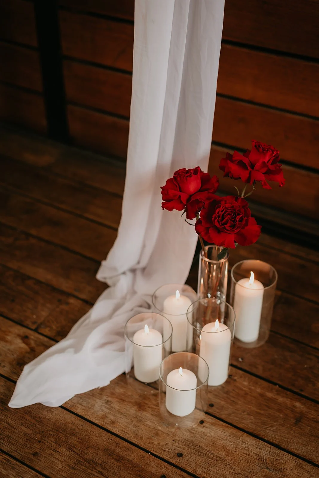Red roses in a clear glass vase, surrounded by lit white candles in glass holders, on a white cloth against a wooden wall.