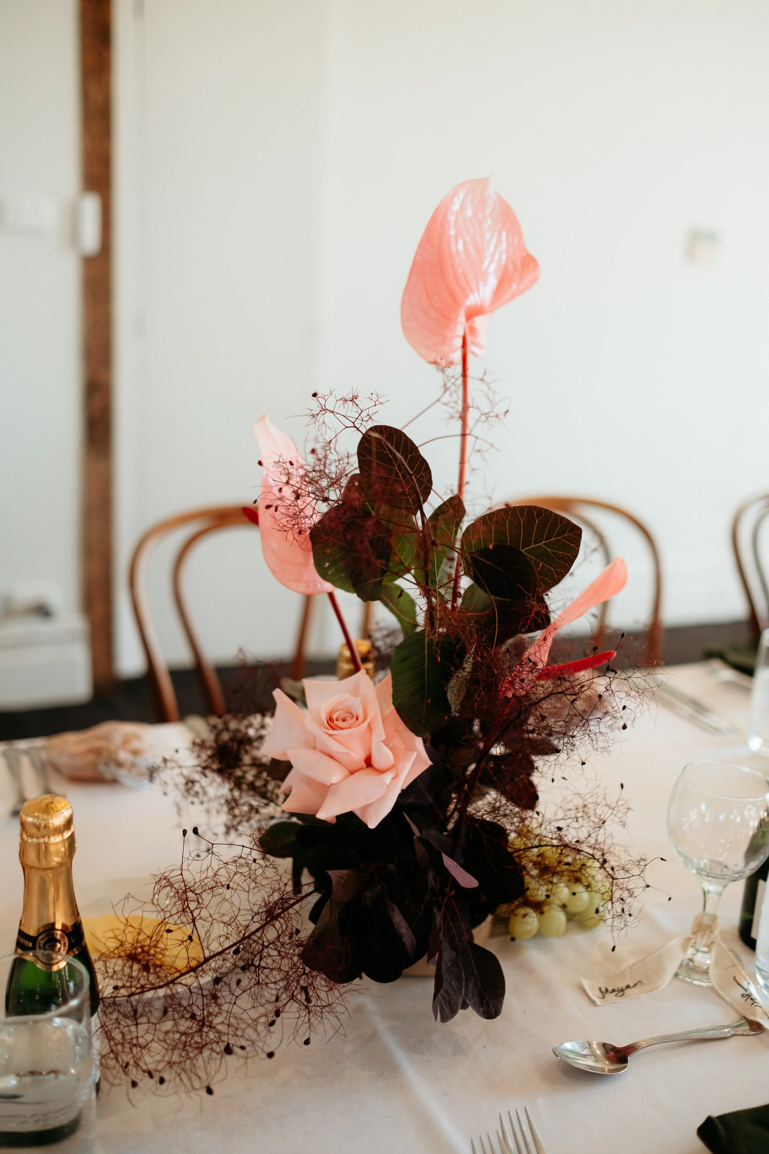 A floral centerpiece with pink roses, anthuriums, dark leaves, and decorative twigs on a dinner table at a formal event.