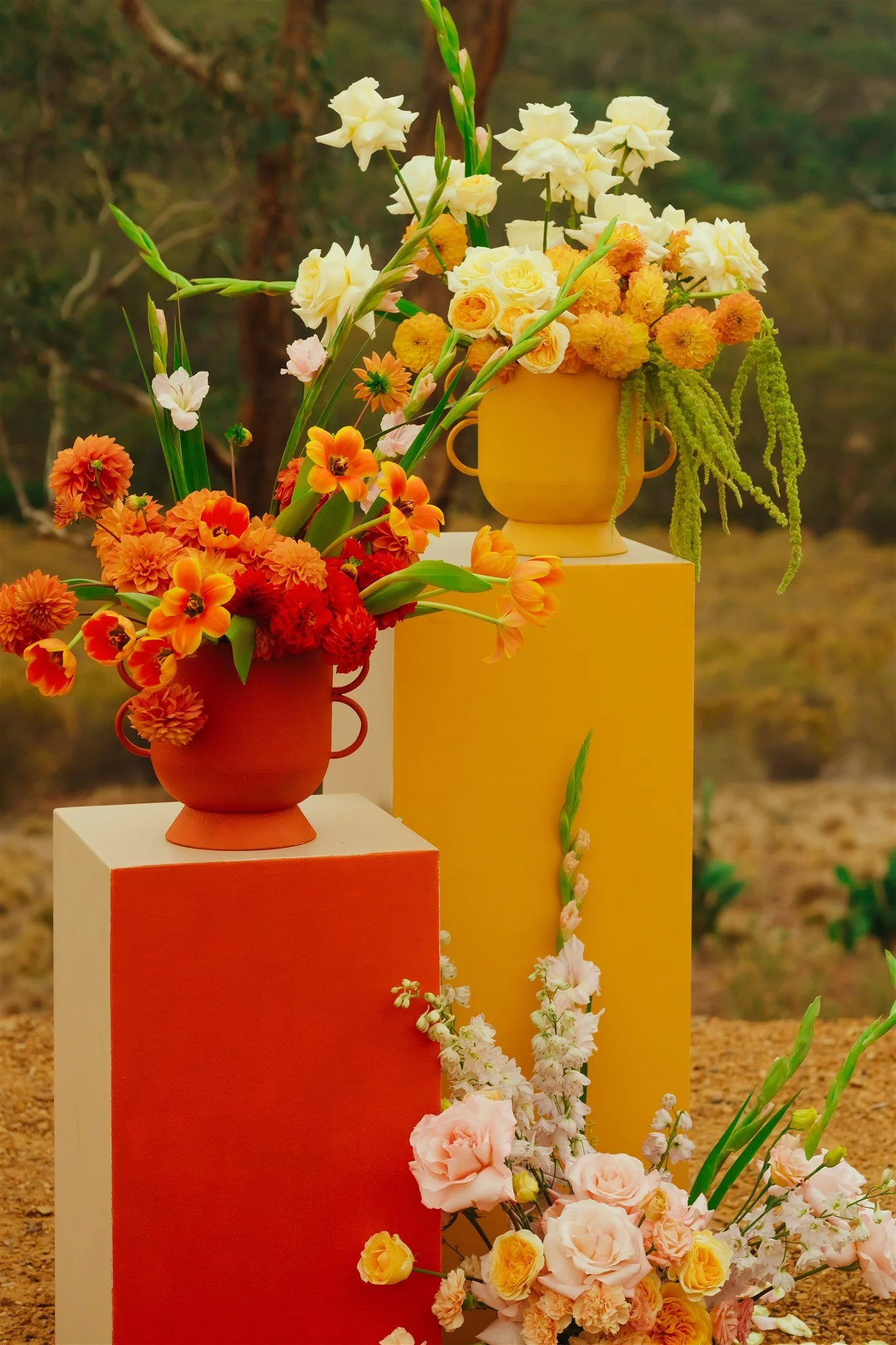 Vases with yellow, orange, and pink flowers on colorful pedestals outdoors.