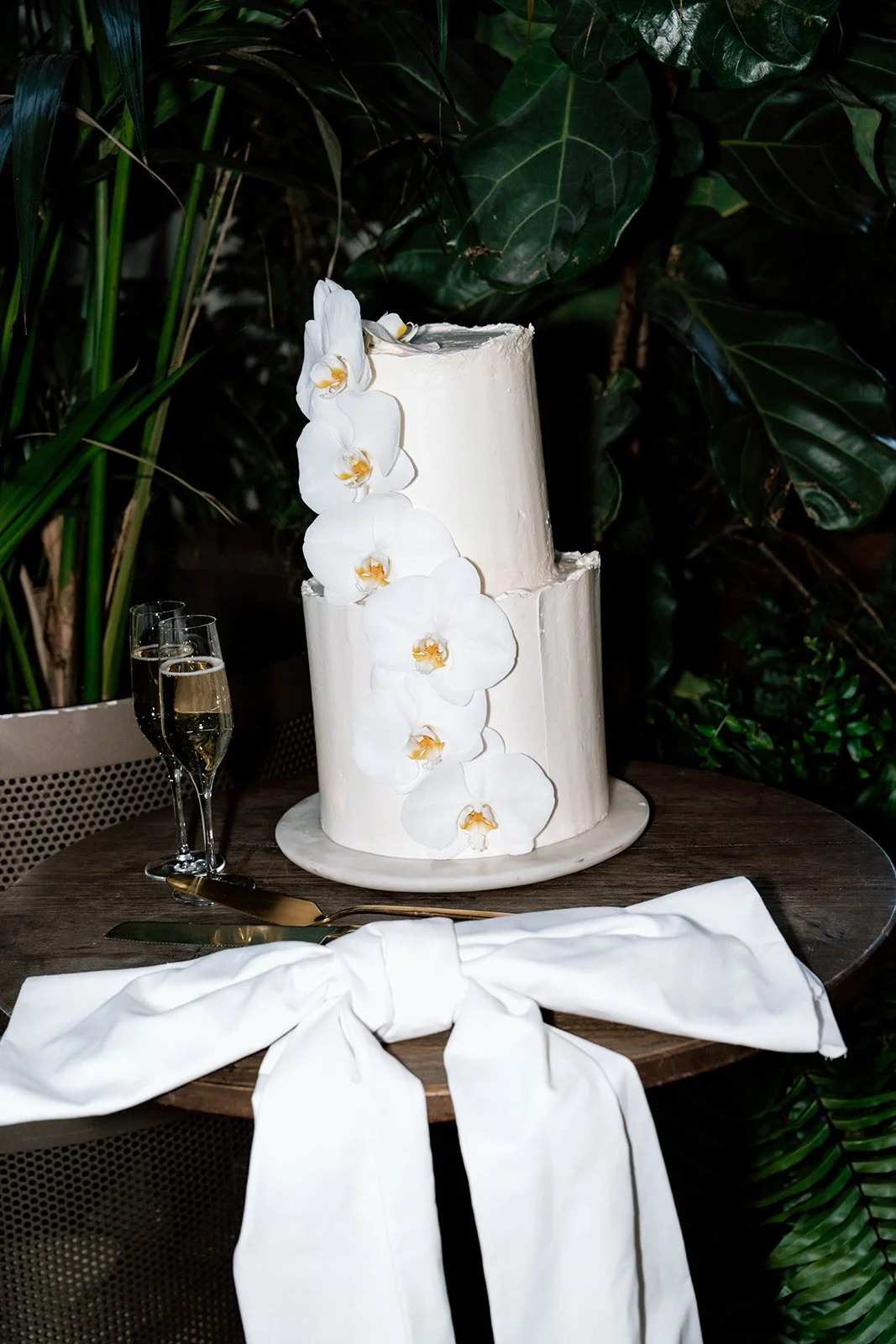 A two-tiered white wedding cake decorated with cascading white orchids, placed on a wooden table. Two glasses of champagne and a white napkin tied in a bow are nearby, with lush green foliage in the background.