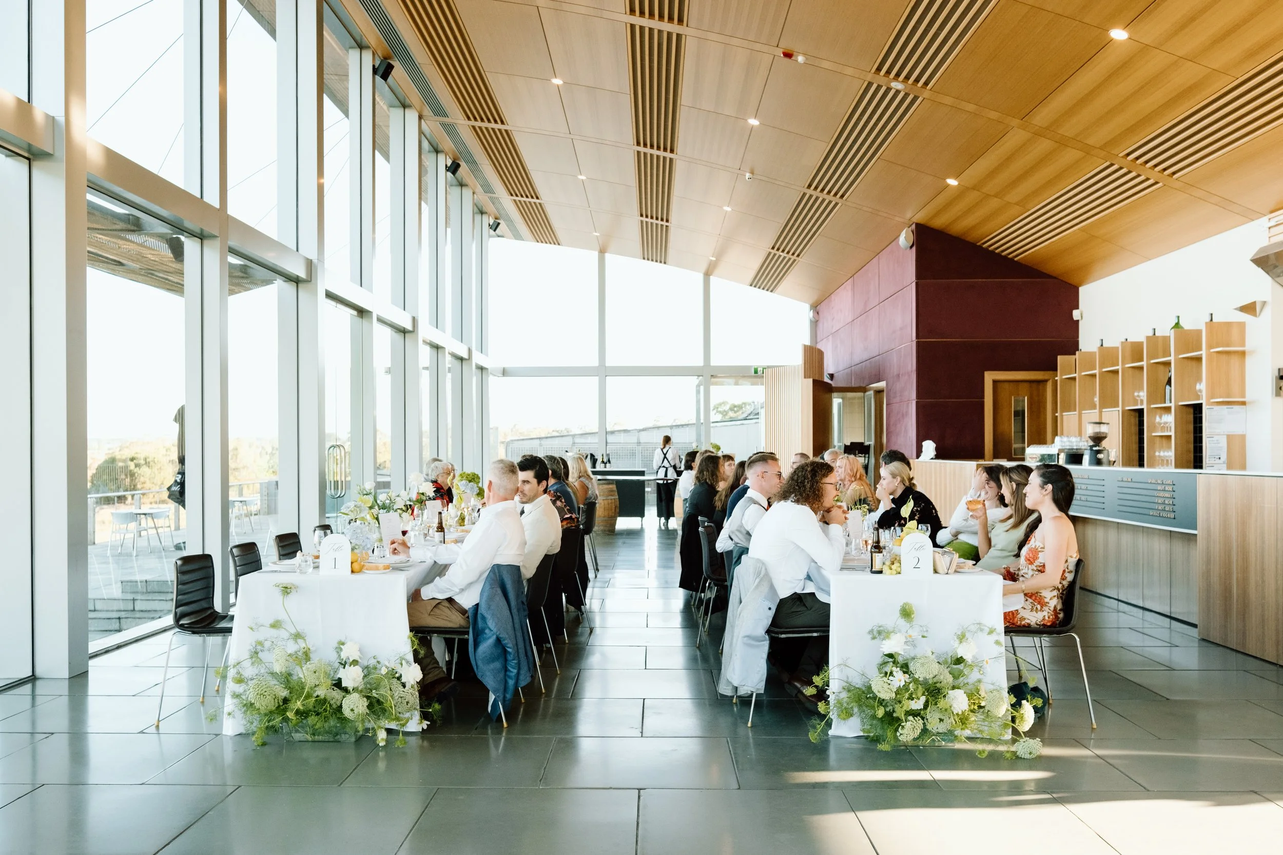 People dining at tables with white tablecloths and floral centerpieces inside a modern building with large windows and wooden ceiling.