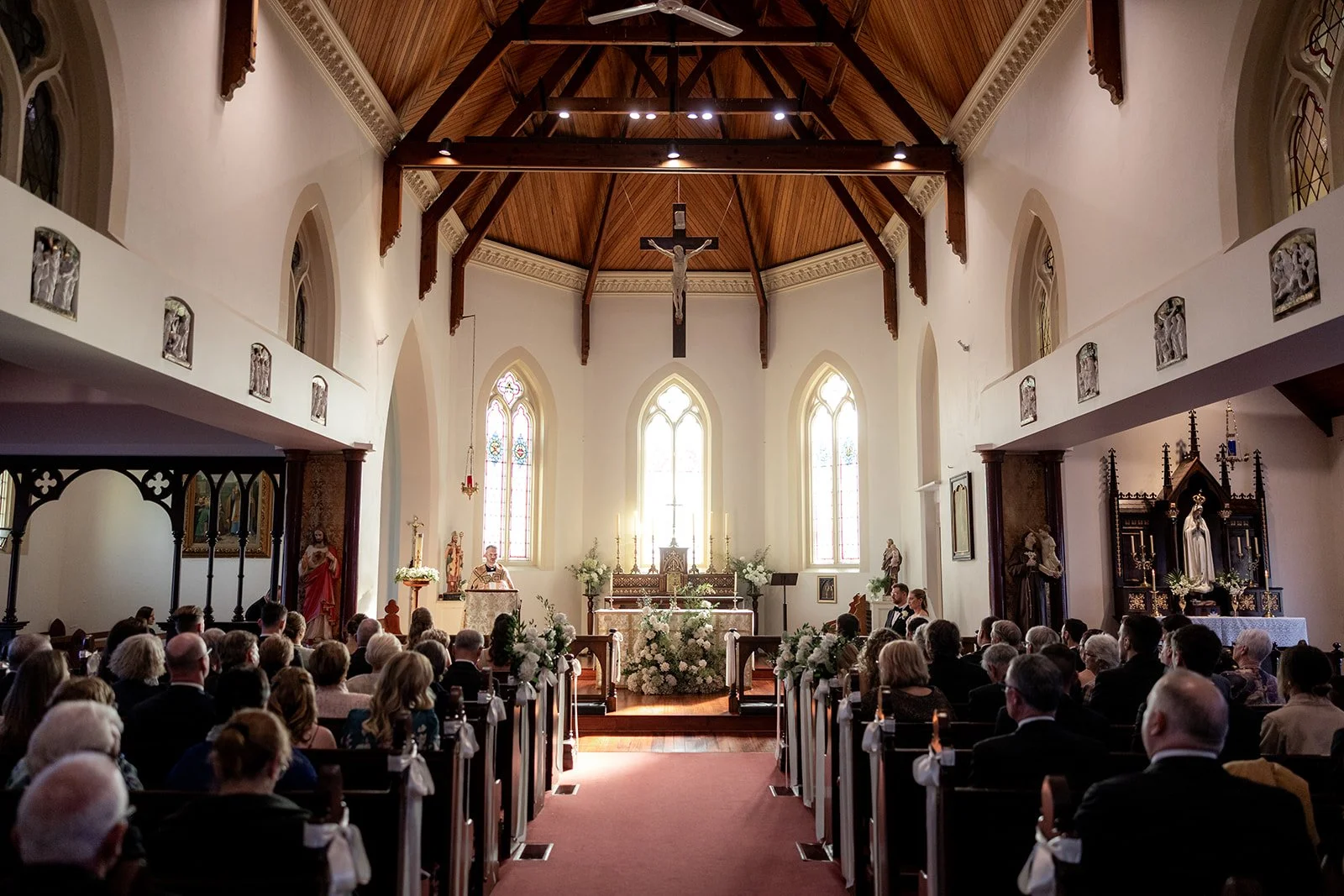 Interior of a church with a congregation attending a wedding ceremony. The altar is decorated with flowers, and a priest is presiding over the event. The church features stained glass windows and wooden ceiling beams.