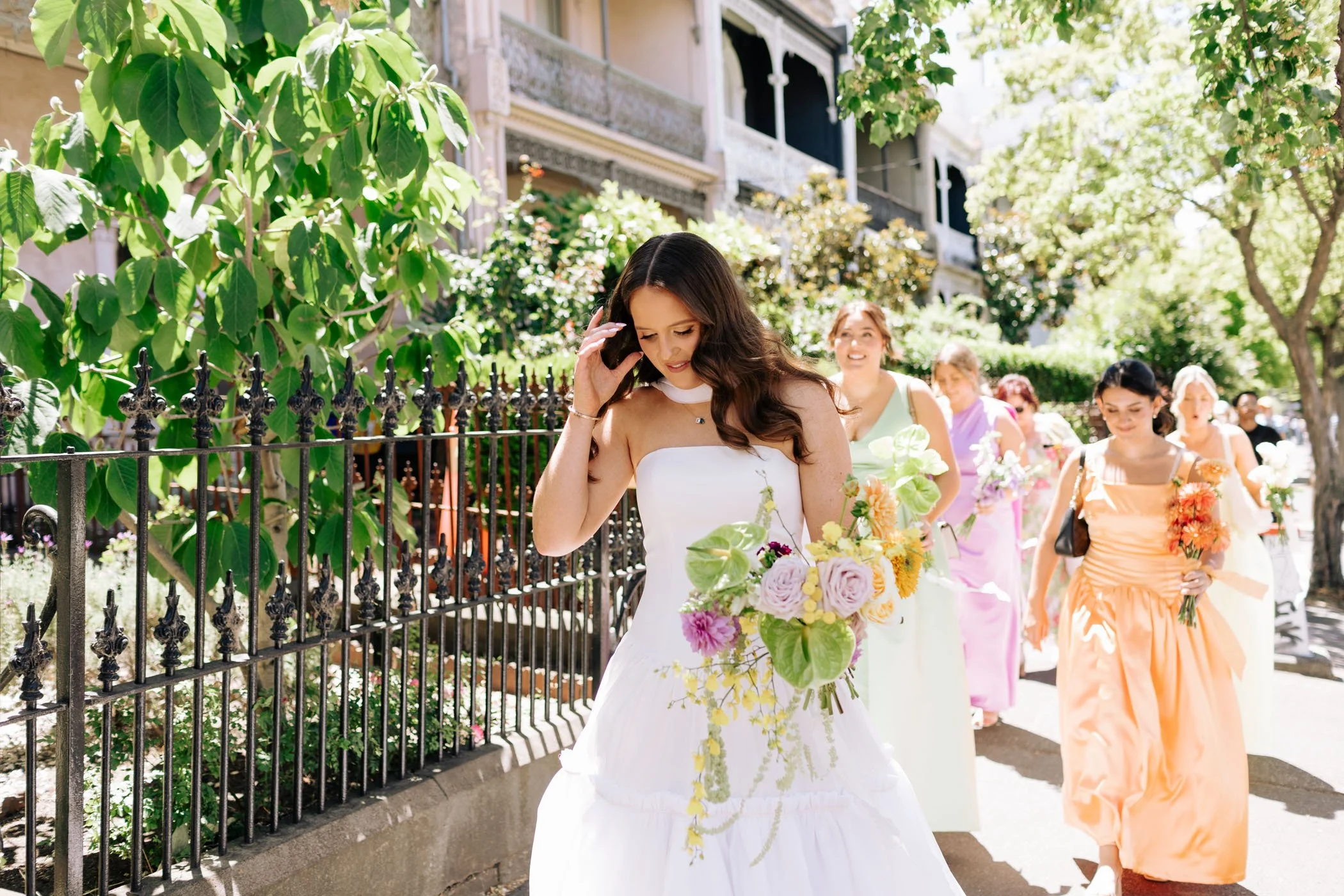 A group of women walking outdoors, with the woman in the foreground holding a bouquet of flowers, wearing a white dress, and smiling. The scene includes trees, greenery, and buildings in the background, indicating a sunny day.