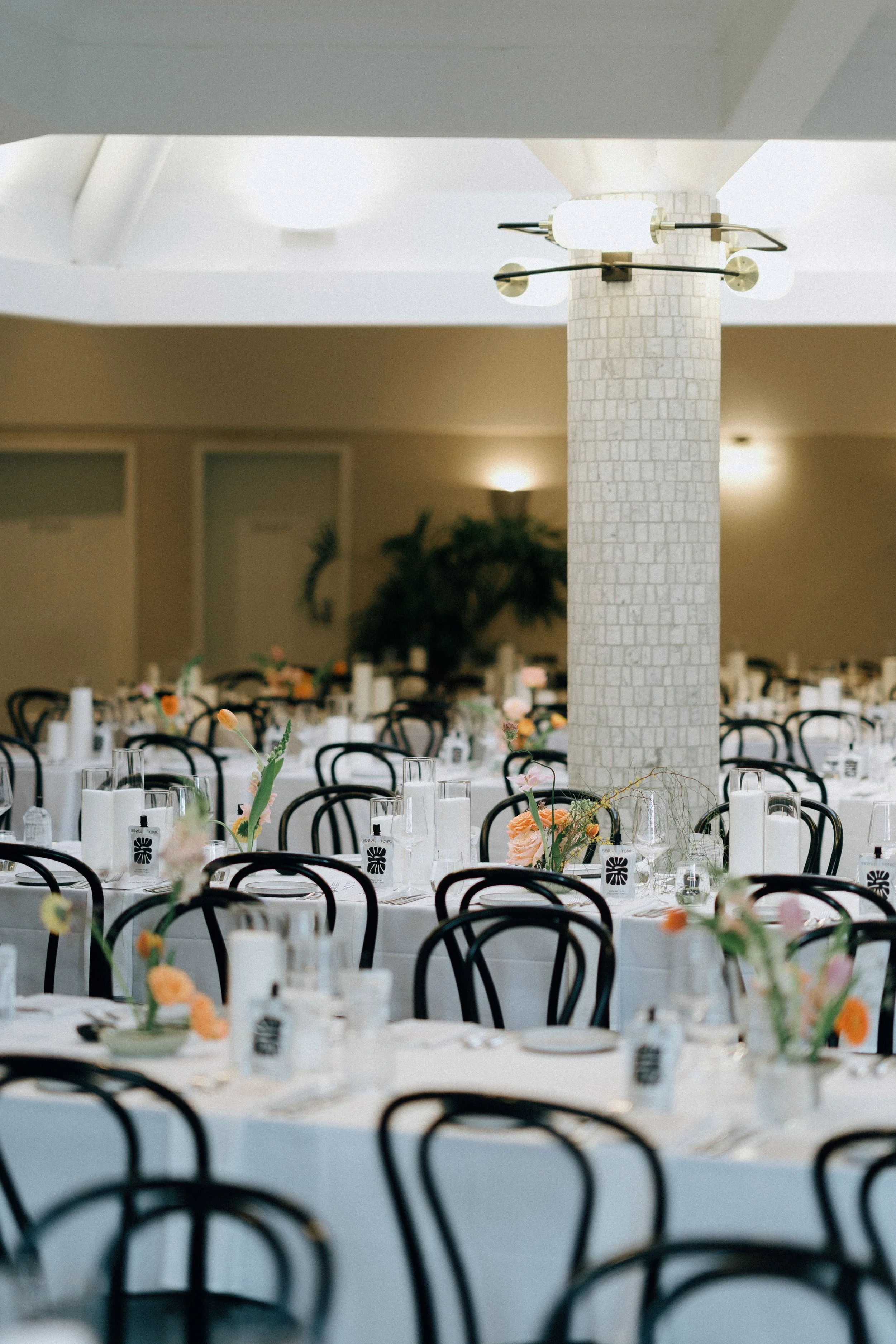 Round tables decorated with floral centerpieces, each with wine glasses and place settings, in a decorated event space with chairs and a large white tiled column.
