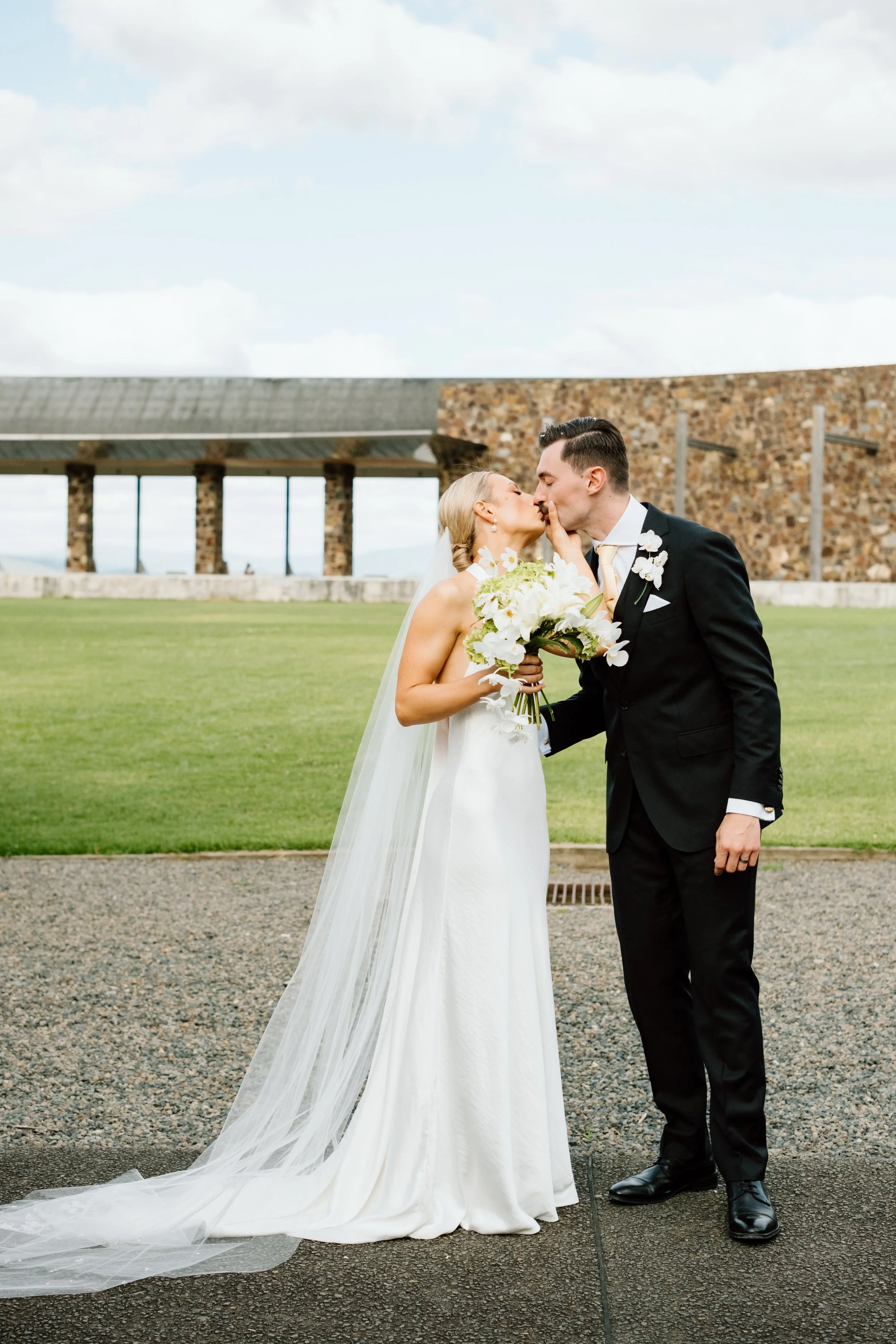 A bride and groom sharing a kiss during their wedding ceremony outdoors on a cloudy day.