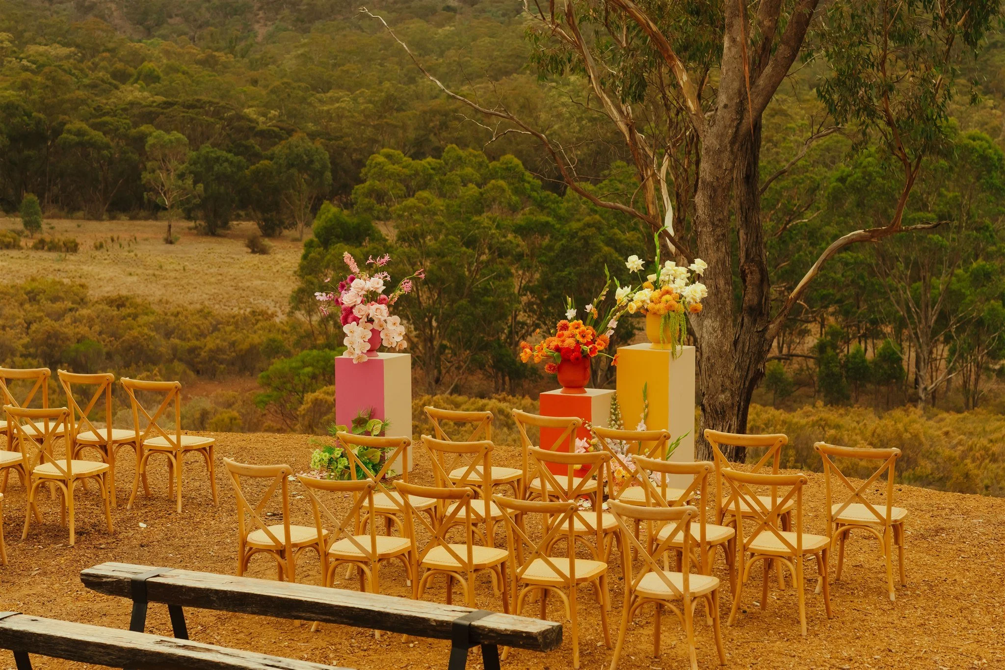 Outdoor setting with rows of wooden chairs and colorful flower arrangements on pedestals, set against a backdrop of rolling hills and trees.