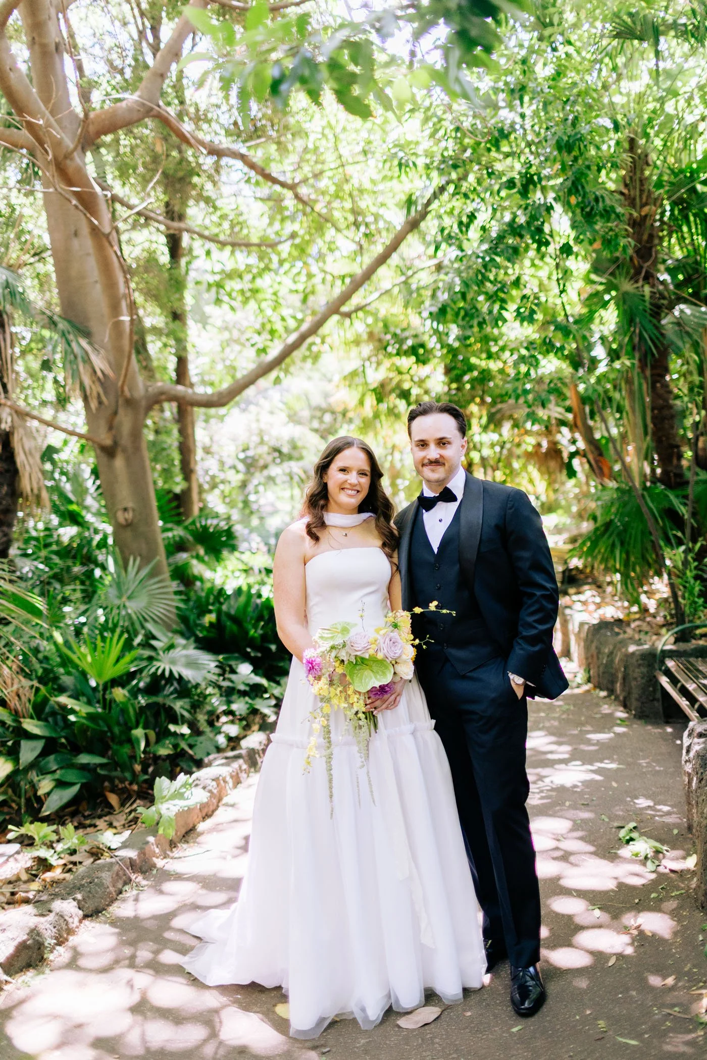 A bride and groom standing together outdoors in a lush, green garden or park. The bride wears a white strapless dress and holds a bouquet of flowers, while the groom wears a black tuxedo with a bow tie. They are smiling and posing for a photo.