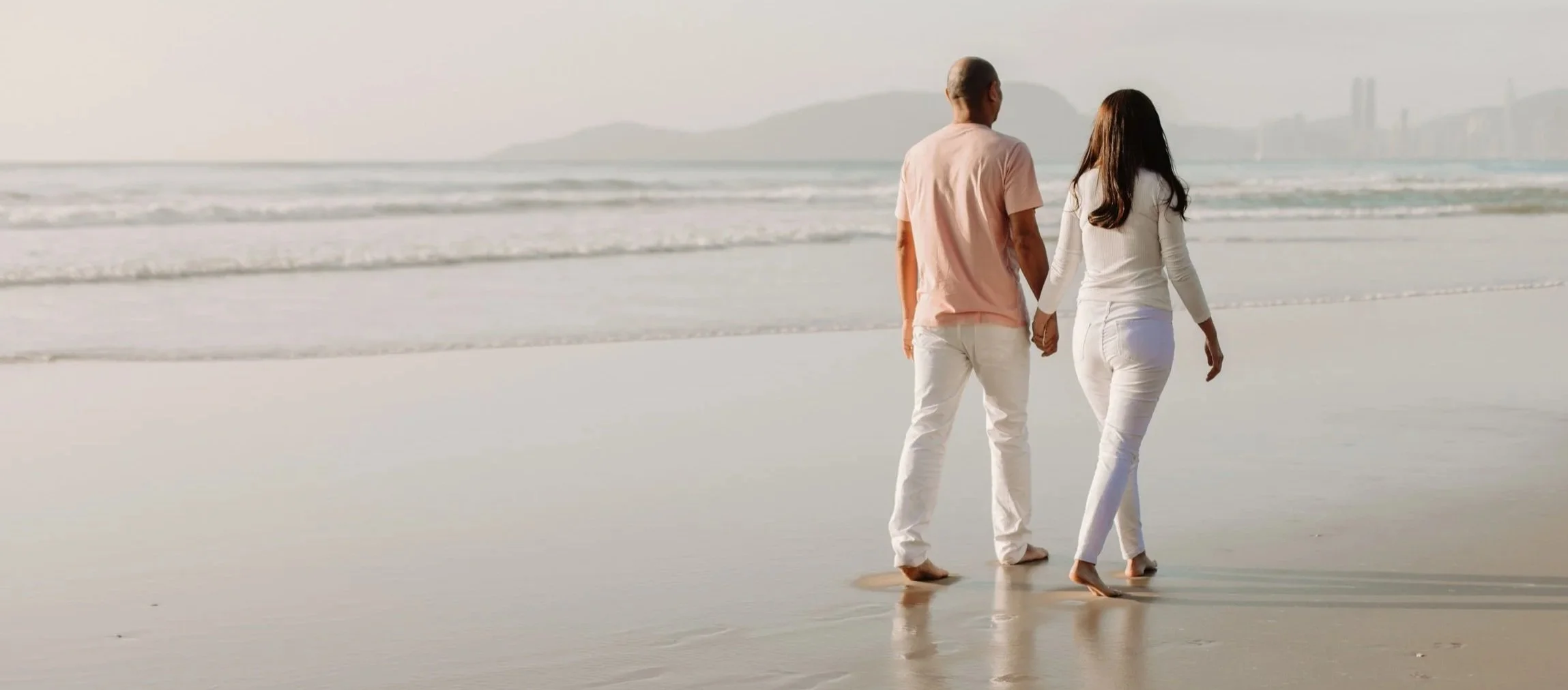 A couple holding hands walking on a sandy beach near the water with a city skyline and mountains in the distance.