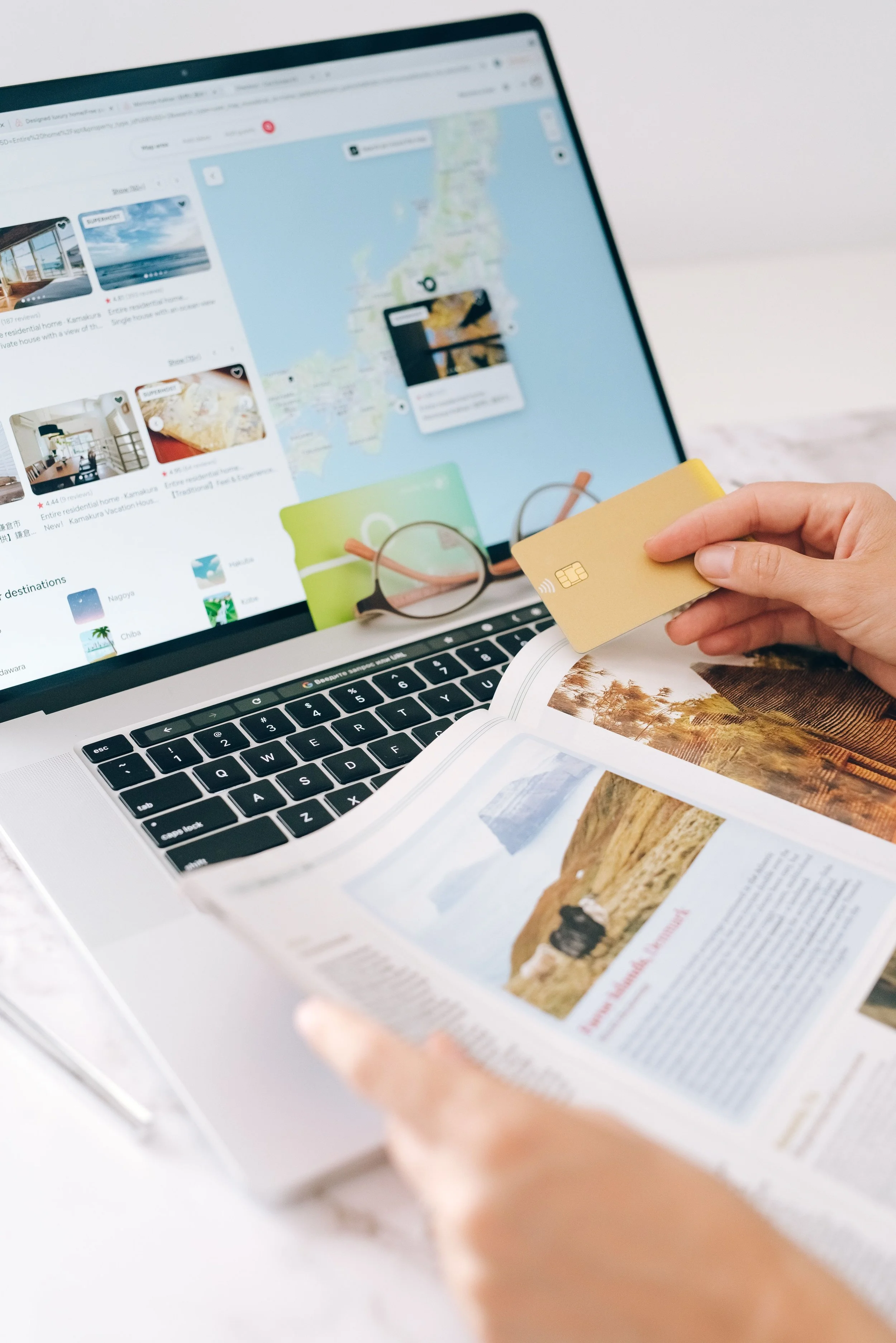 A person holding a gold credit card next to eyeglasses and an open magazine on a white surface, with a laptop in the background displaying a map and vacation listings.