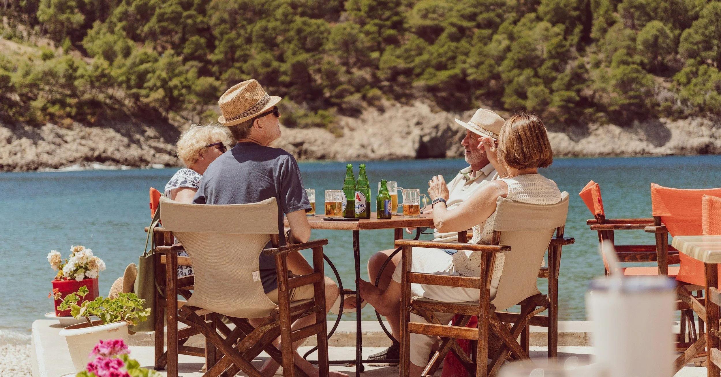 Group of four elderly people sitting at a table outdoors by a lake, enjoying drinks and conversation on a sunny day, with green trees on the hillside in the background.