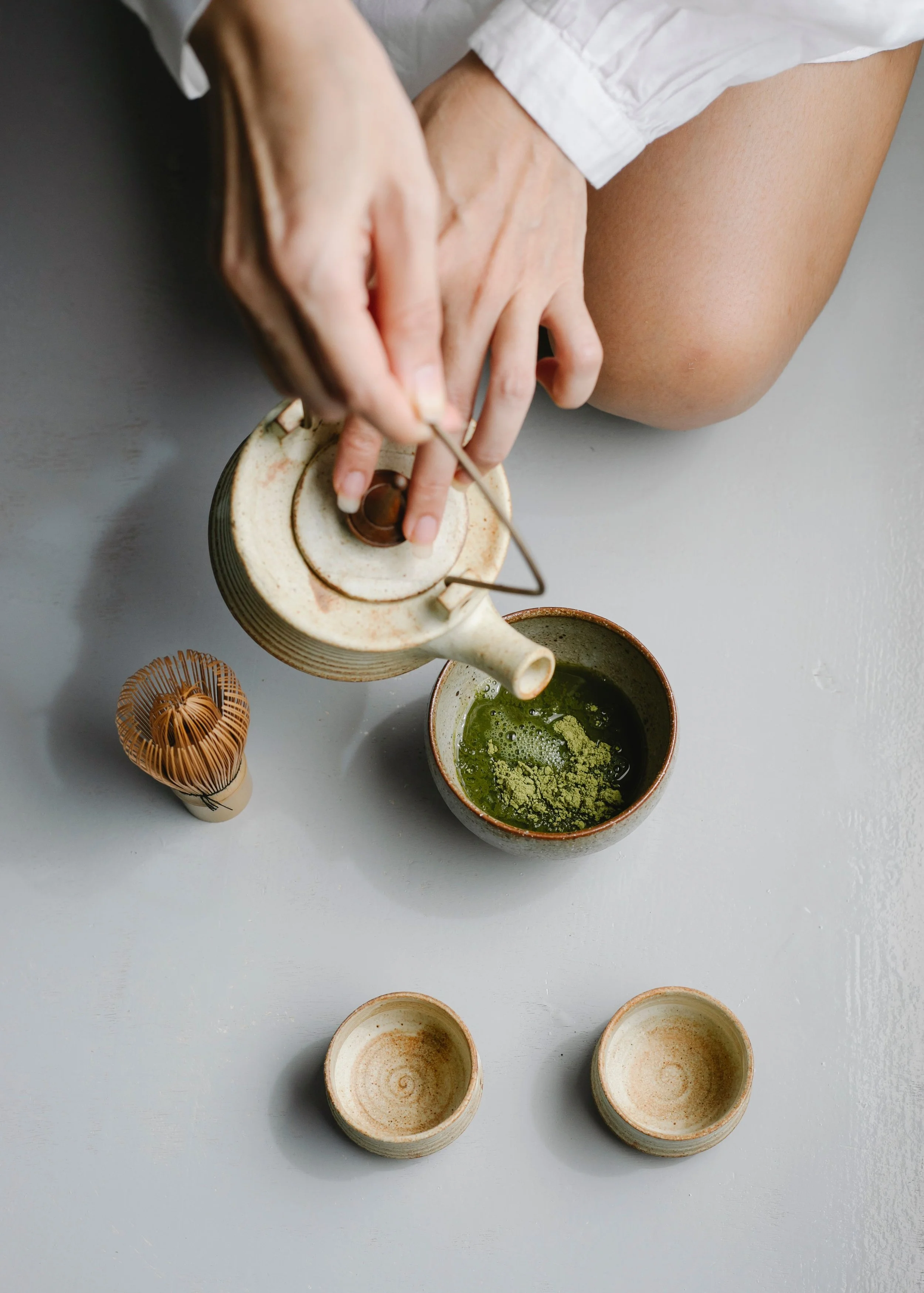 A person is preparing matcha tea using a traditional whistling kettle and a bowl of green matcha powder, with empty tea bowls and tea accessories on a light gray surface.