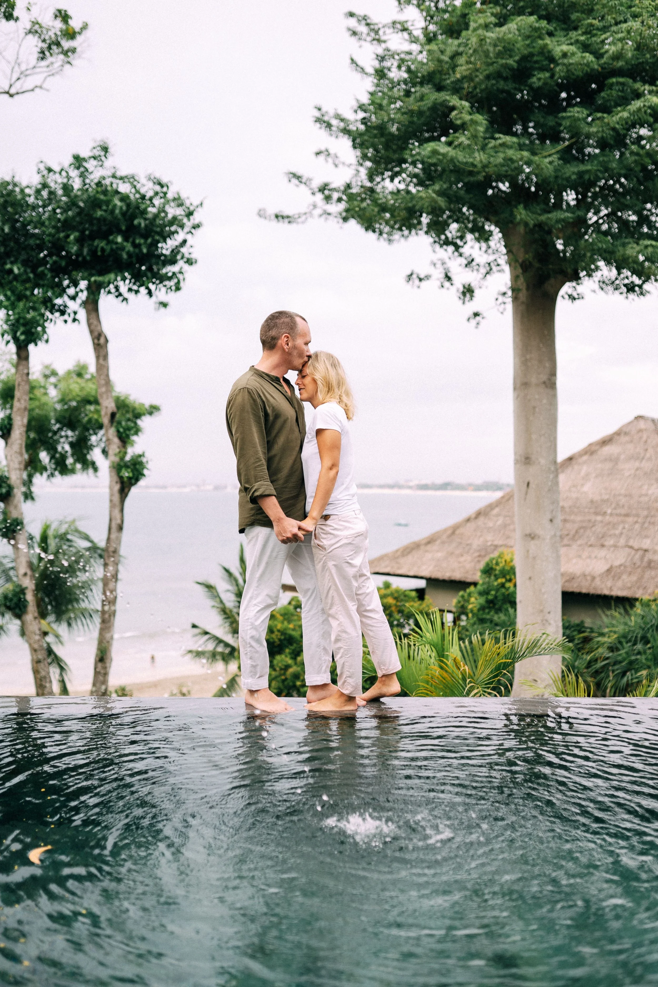 A couple holding hands and touching foreheads, standing barefoot in a pool with a scenic background of trees, a lake, and a thatched-roof structure, under an overcast sky.