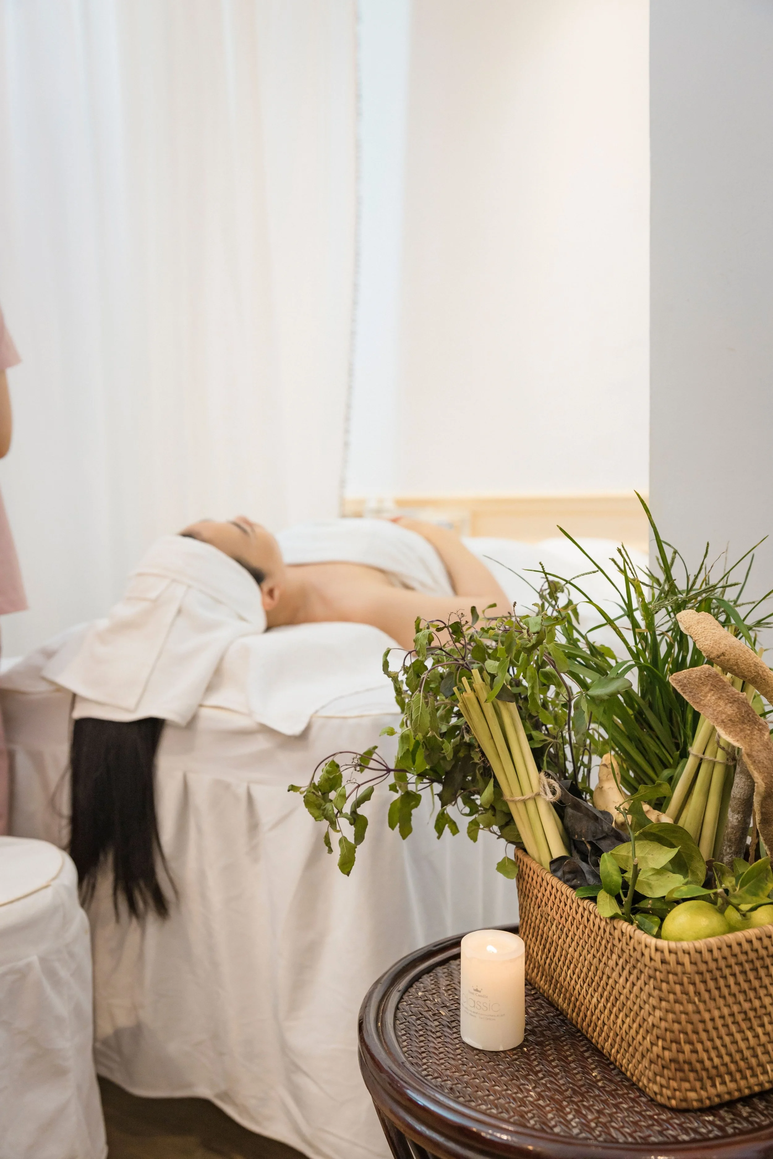 Person lying on a massage table with a towel wrapped around their head, in a spa or massage room with white curtains and a wicker basket of green plants in the foreground.