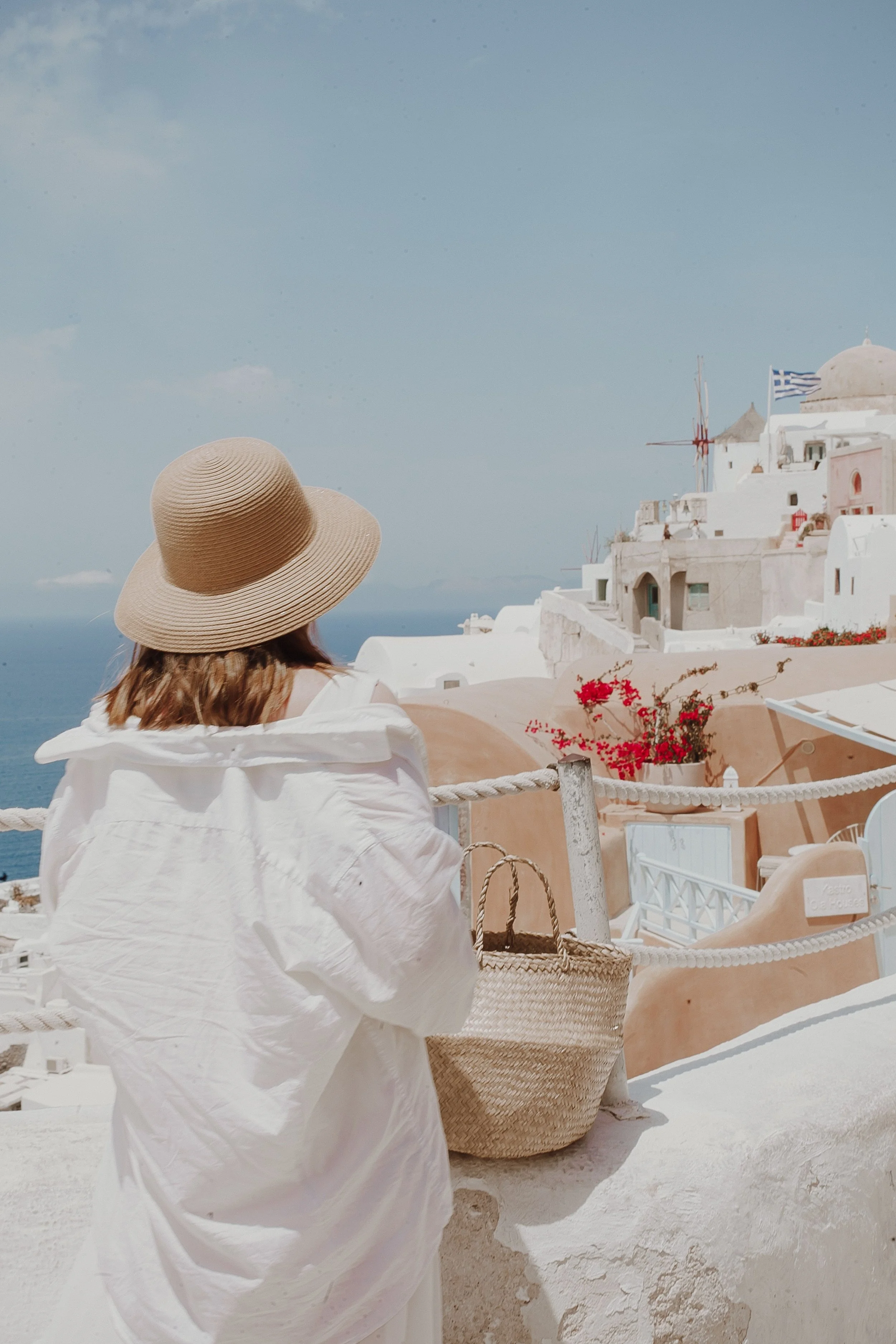 A woman wearing a wide-brimmed straw hat and a white dress stands on a white terrace overlooking Greek island architecture with white buildings, some with domed roofs and a Greek flag flying, under a partly cloudy sky.