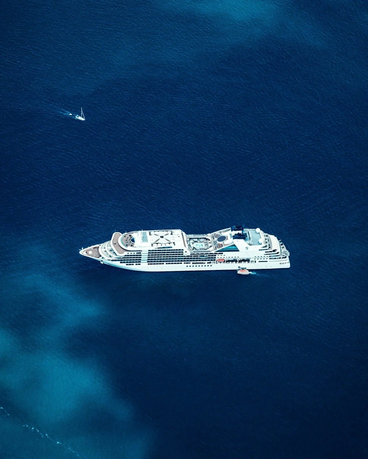 Aerial view of a large white cruise ship sailing on deep blue water with a small sailboat nearby.