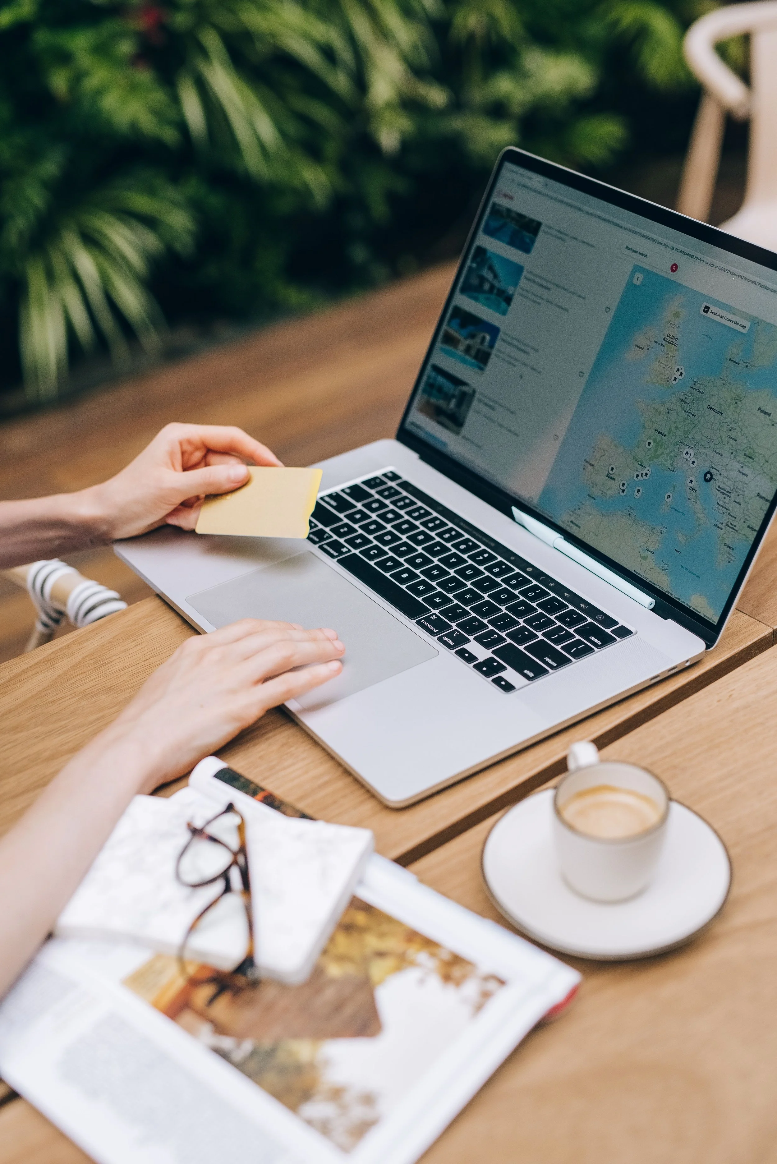 A person holds a yellow sticky note while sitting at a wooden table with a laptop showing a map and images, a cup of coffee, an open magazine, and glasses.