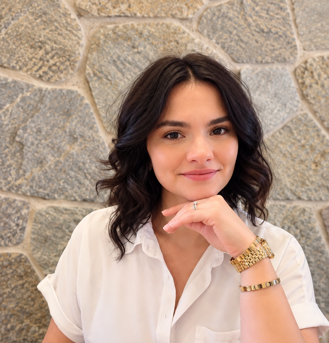 A woman with shoulder-length dark hair and a light skin tone, wearing a white blouse and gold jewelry, sitting in front of a stone wall, looking at the camera with a slight smile.