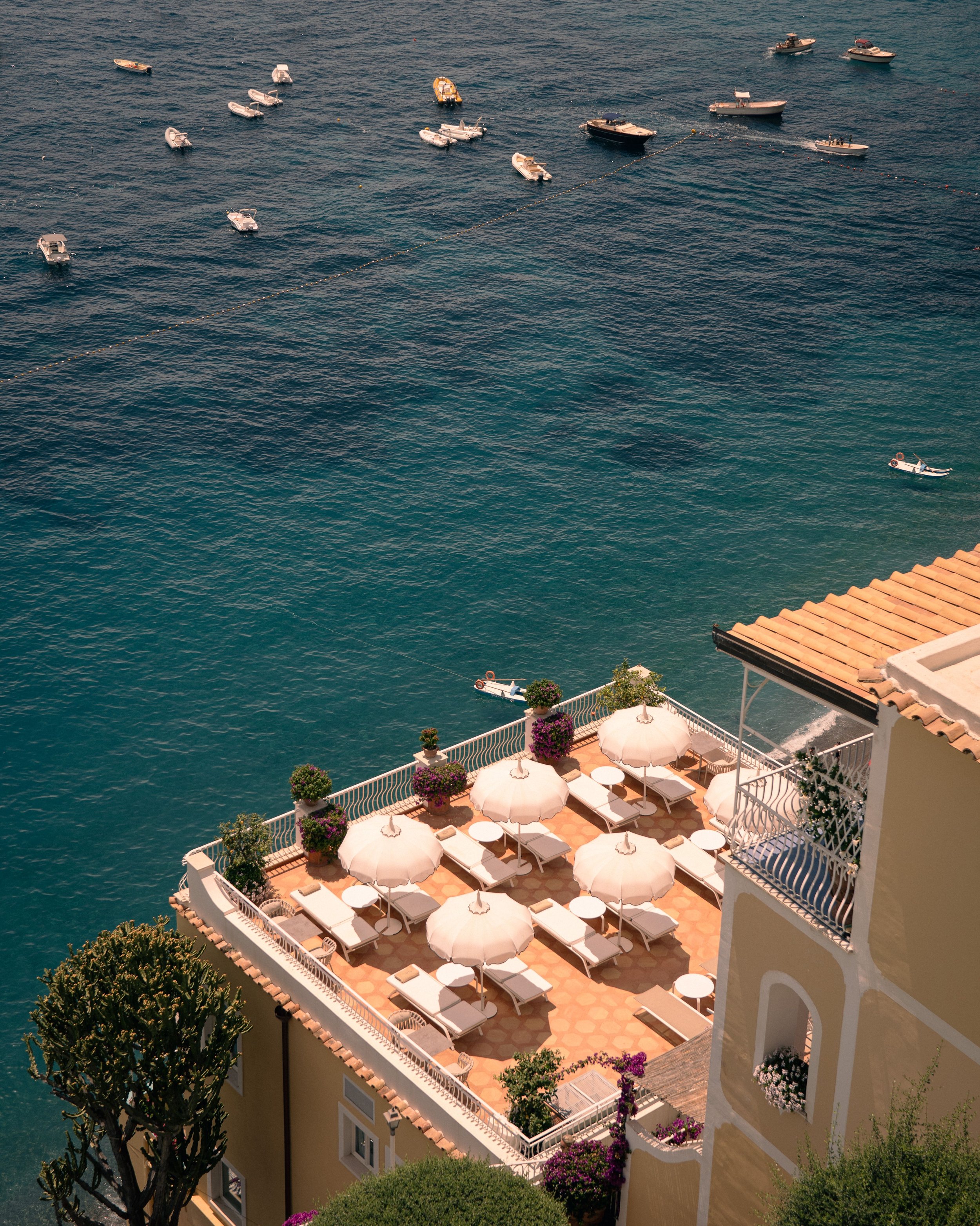 A balcony with white umbrellas and lounge chairs overlooking the ocean with boats floating on the water.