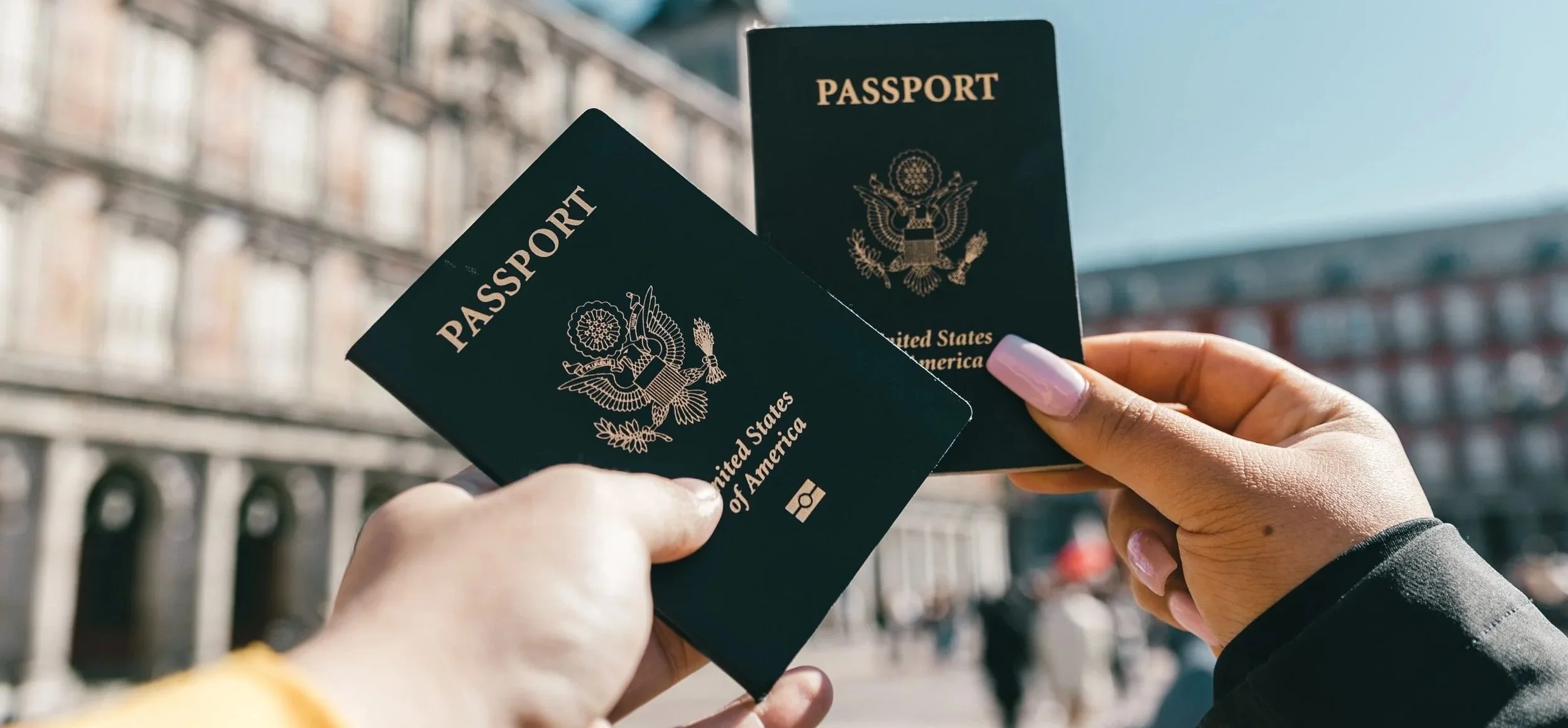 Two people are holding U.S. passports in an outdoor setting with buildings and a clear sky in the background.