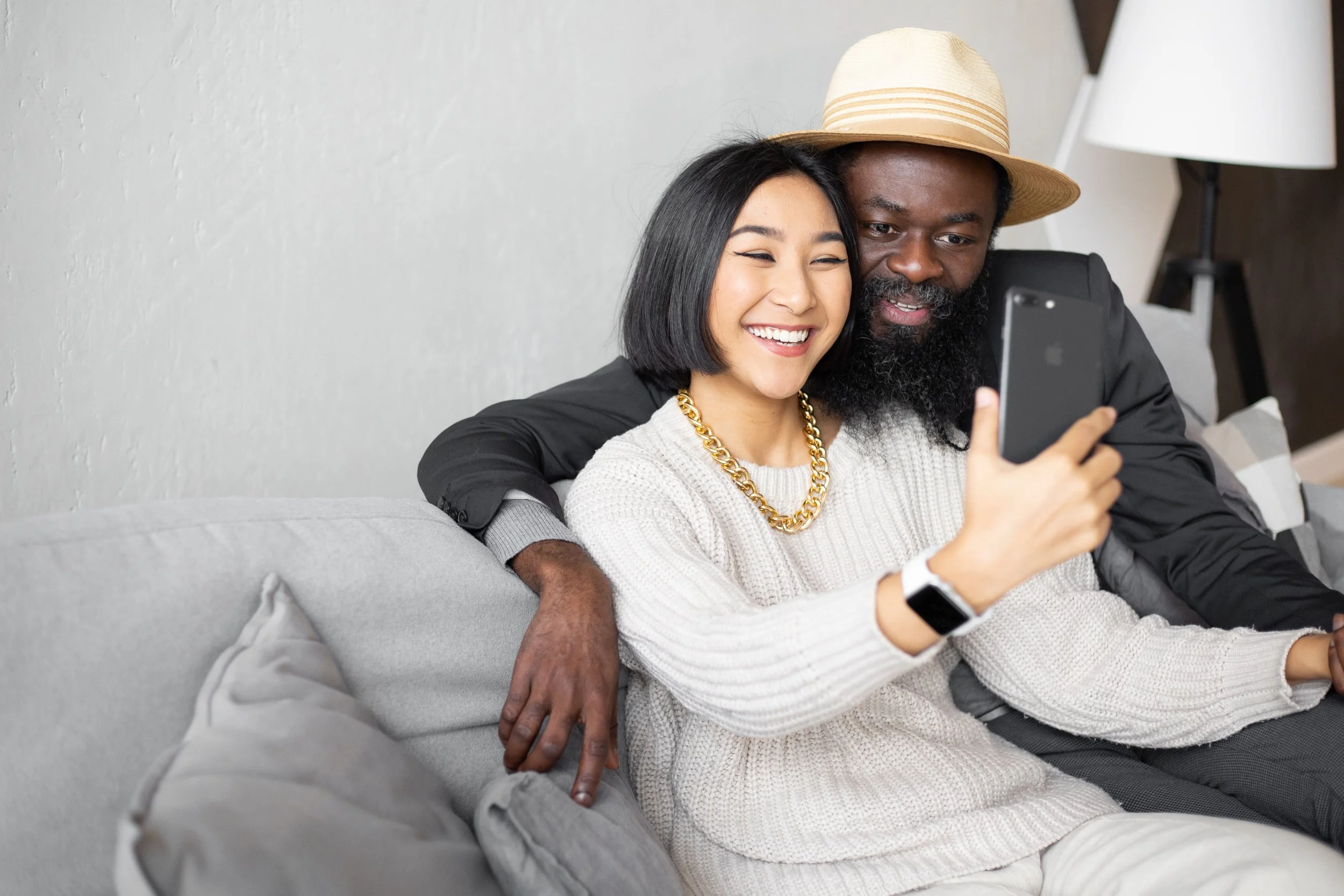 Two people sitting on a light gray couch taking a selfie together. The woman has short black hair, is smiling, and wearing a white sweater with a gold chain necklace. The man has a beard, is wearing a beige straw hat and a black jacket. He is looking at the phone screen with a slight smile. There is a white wall with a lamp in the background.