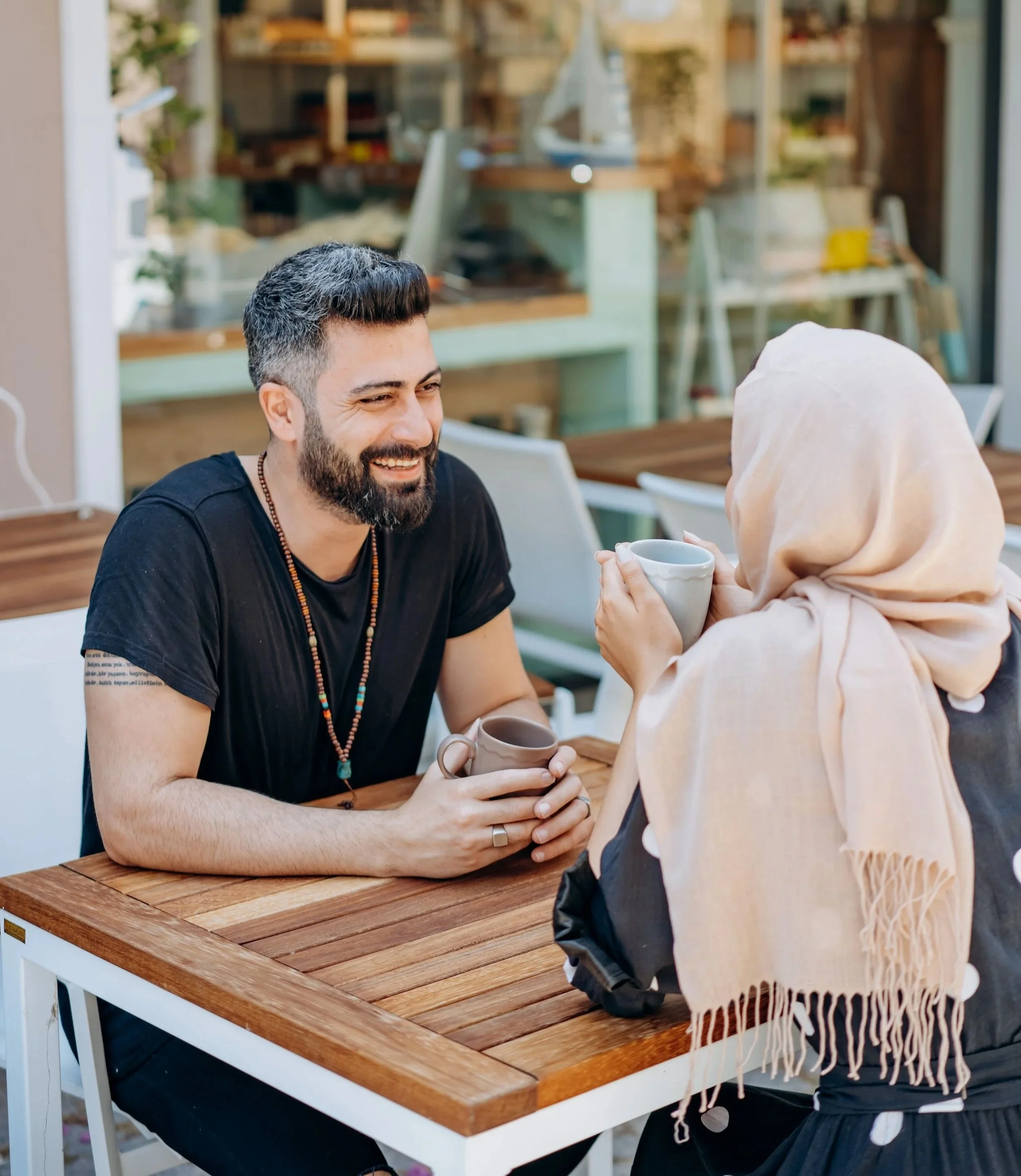 A man with dark hair and beard smiling while sitting at a wooden outdoor table, holding a coffee mug, engaged in conversation with a woman wearing a beige headscarf, in a cozy cafe setting.