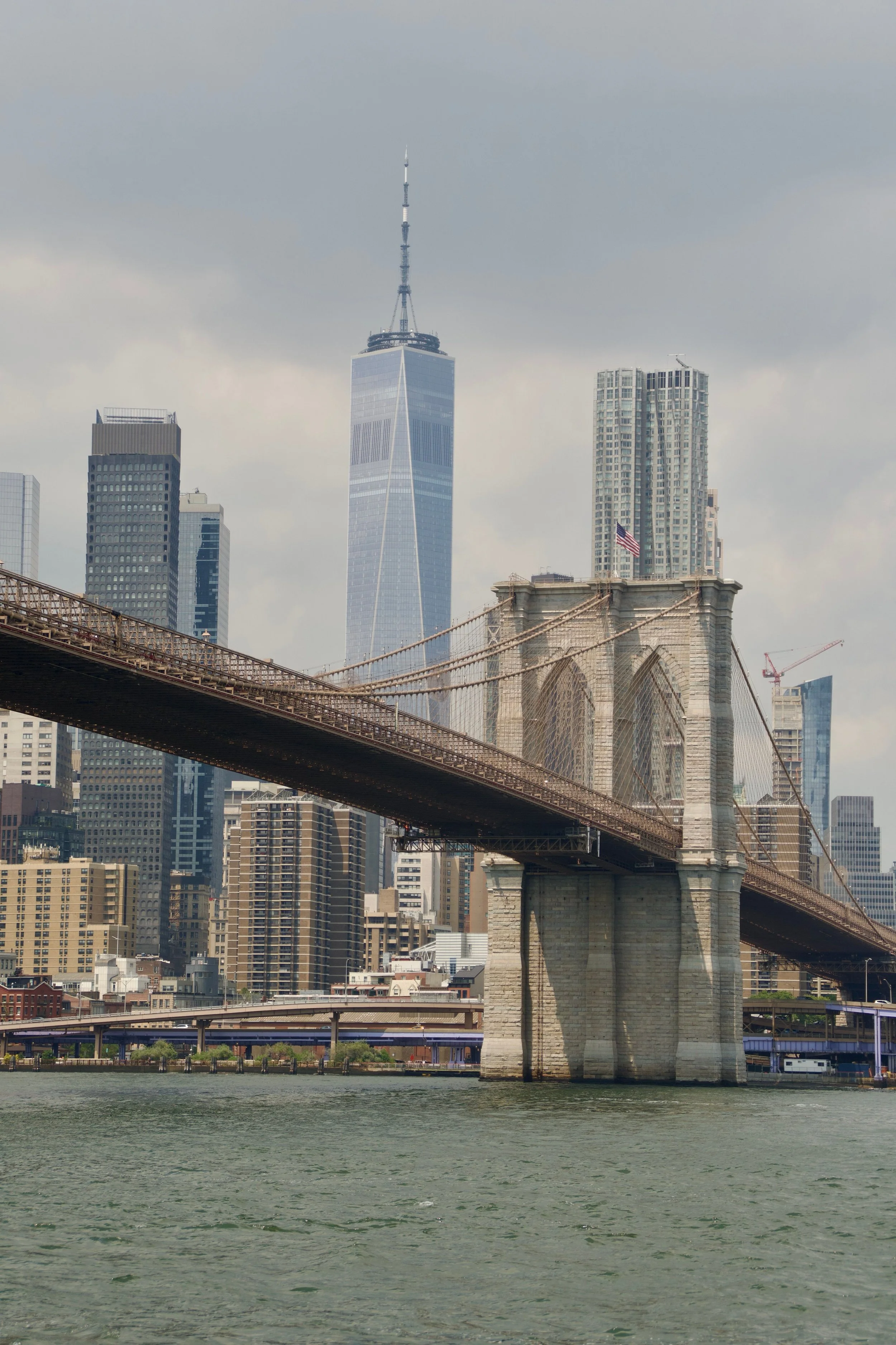 View of the Brooklyn Bridge with the One World Trade Center in Manhattan, New York City, visible behind it, under a partly cloudy sky.