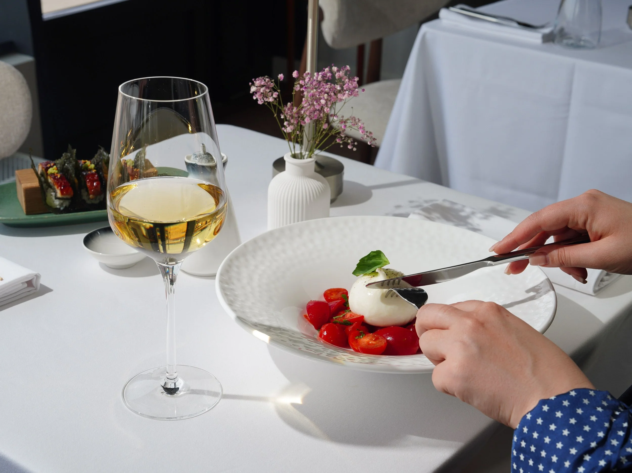 A glass of white wine on a table with a small white bowl of salad, a plate of sushi, a vase with pink flowers, and a person cutting a salad in a white bowl.